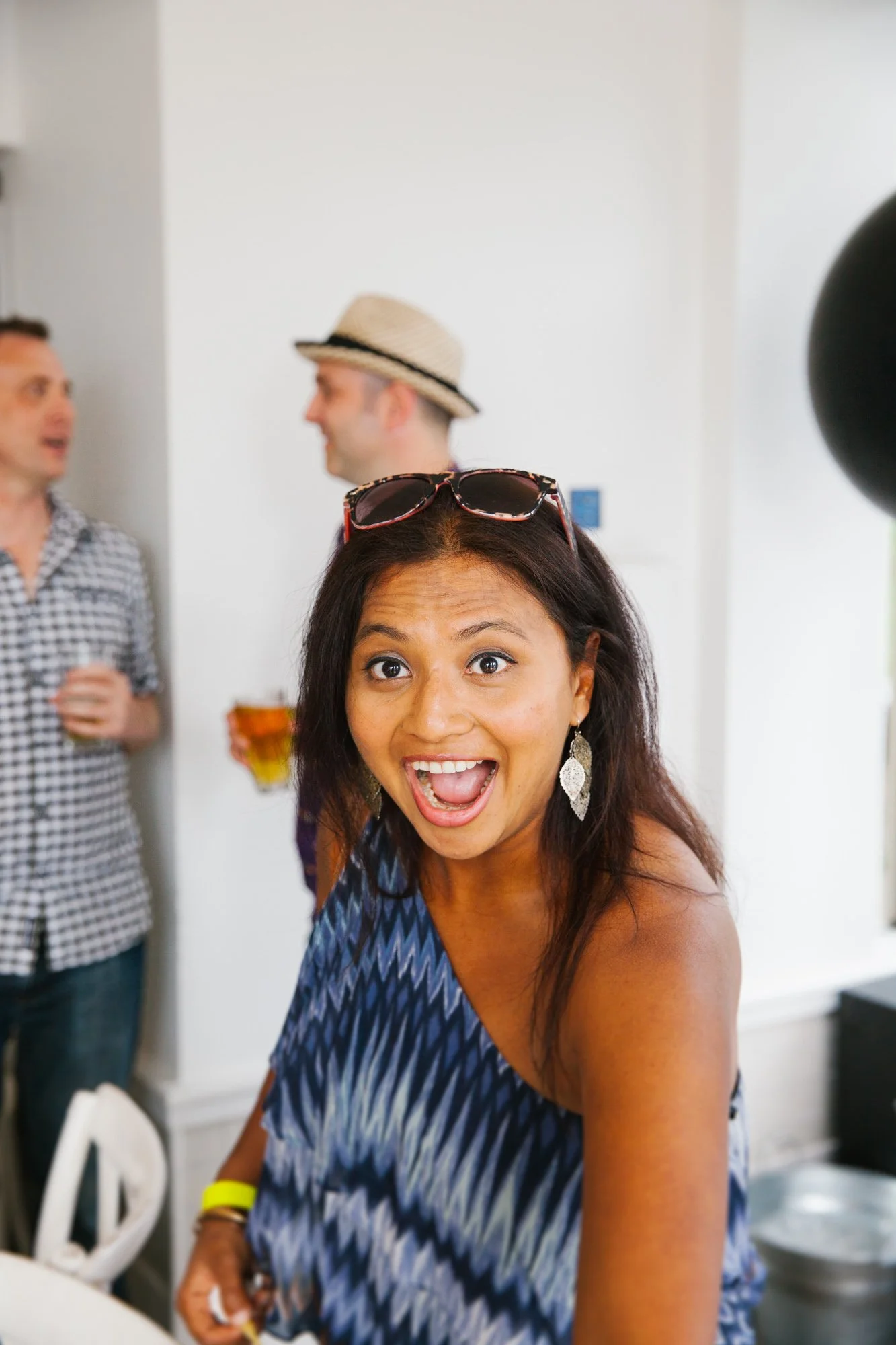 A woman with long dark hair, wearing a blue patterned sleeveless top and sunglasses on her head, smiling with an open mouth. Two men are in the background in a social setting, one wearing a checkered shirt and the other wearing a straw hat, holding b