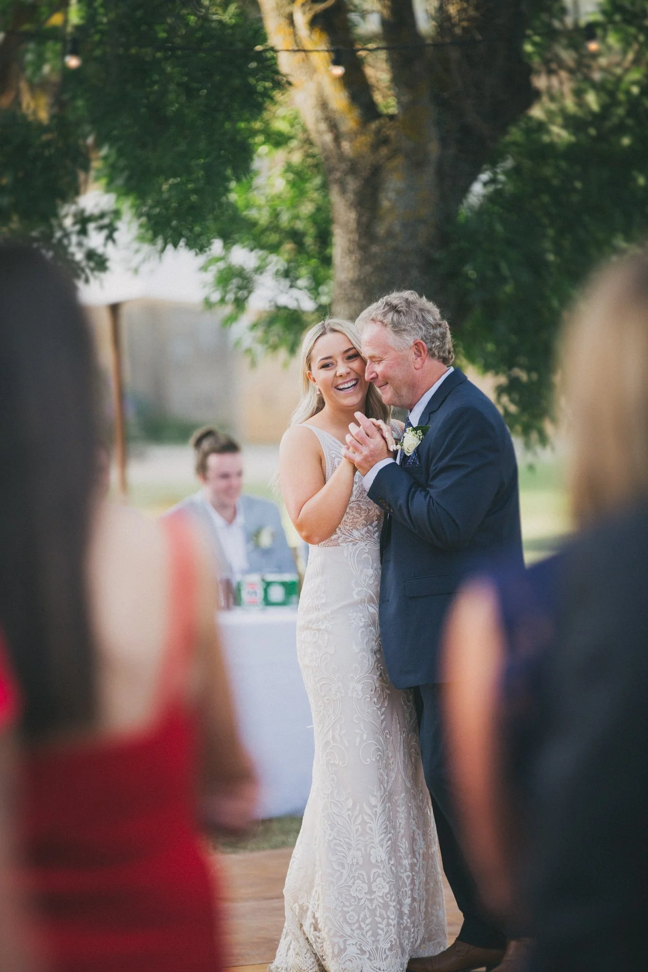 A bride and an older man, possibly her father, sharing a dance at an outdoor wedding reception under a large tree.