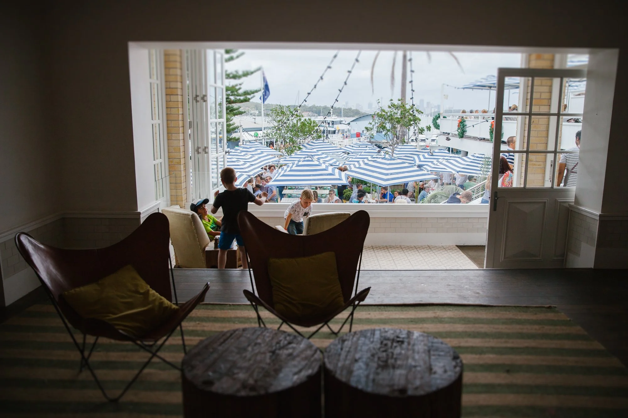 View from inside a room looking out onto a crowded outdoor patio with blue and white striped umbrellas, trees, and many people.