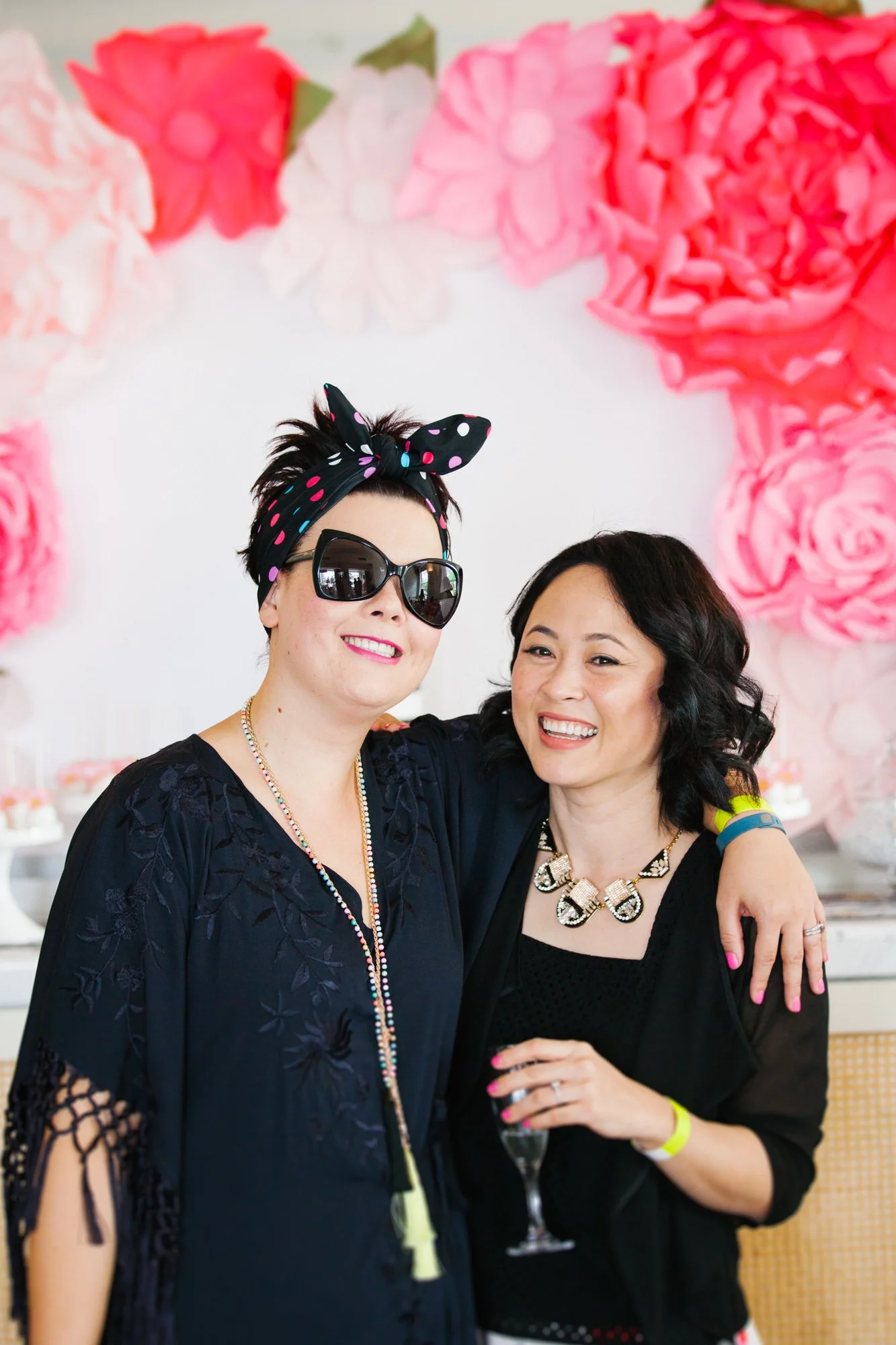 Two women smiling and celebrating at a festive event, with pink and white paper flower decorations in the background. One woman is wearing sunglasses, a polka dot headscarf, and a black lace dress, while the other is wearing a black top and a stateme