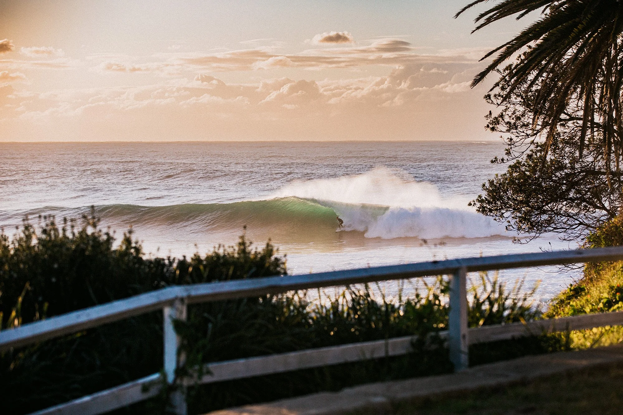 A surfer riding a wave near the shore during sunset, with boats and clouds in the background, trees on the right, and a white fence in the foreground.