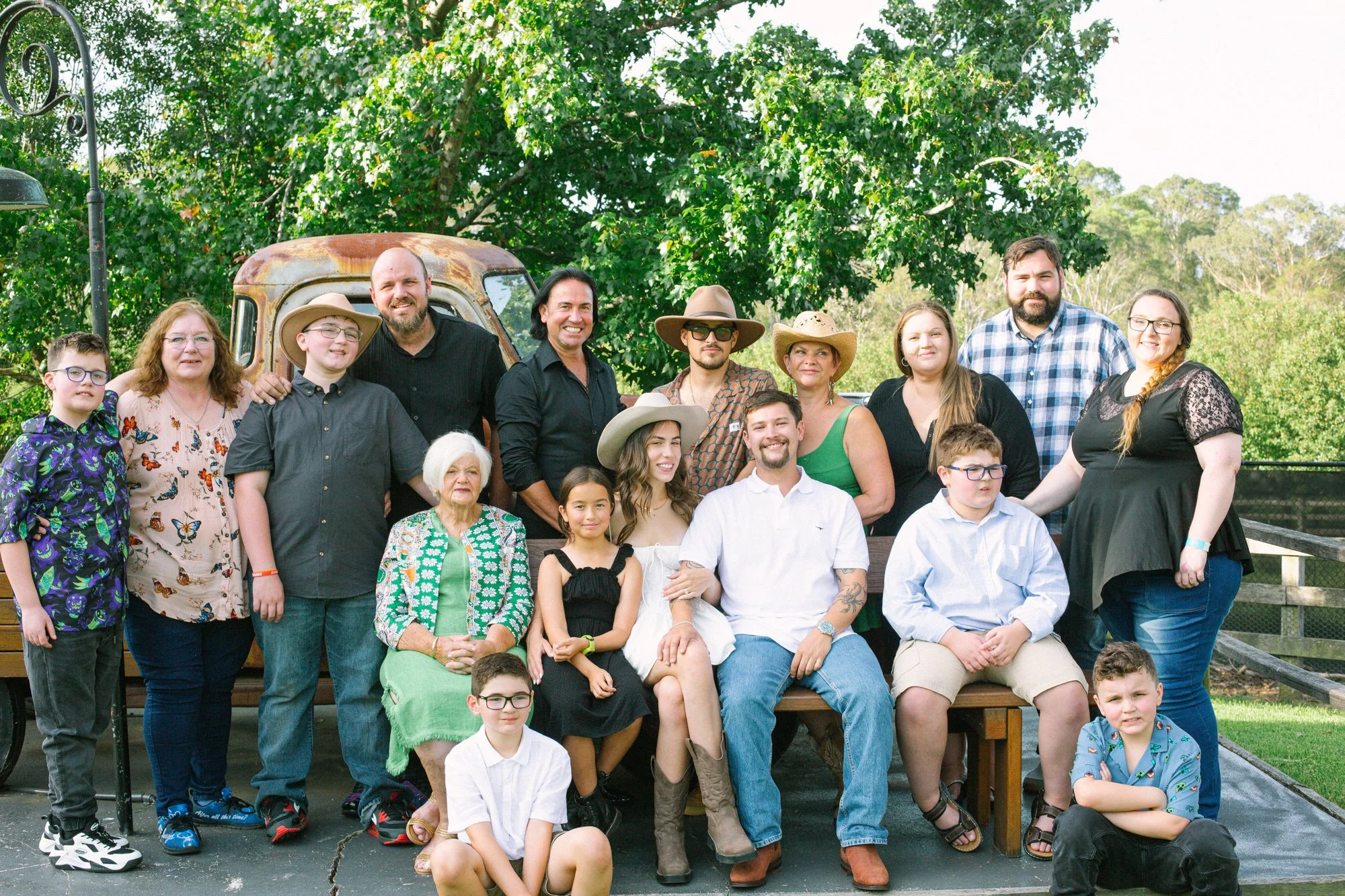 A large group of people posing together outdoors with trees and a rusty vehicle in the background, some wearing hats and casual summer clothing.
