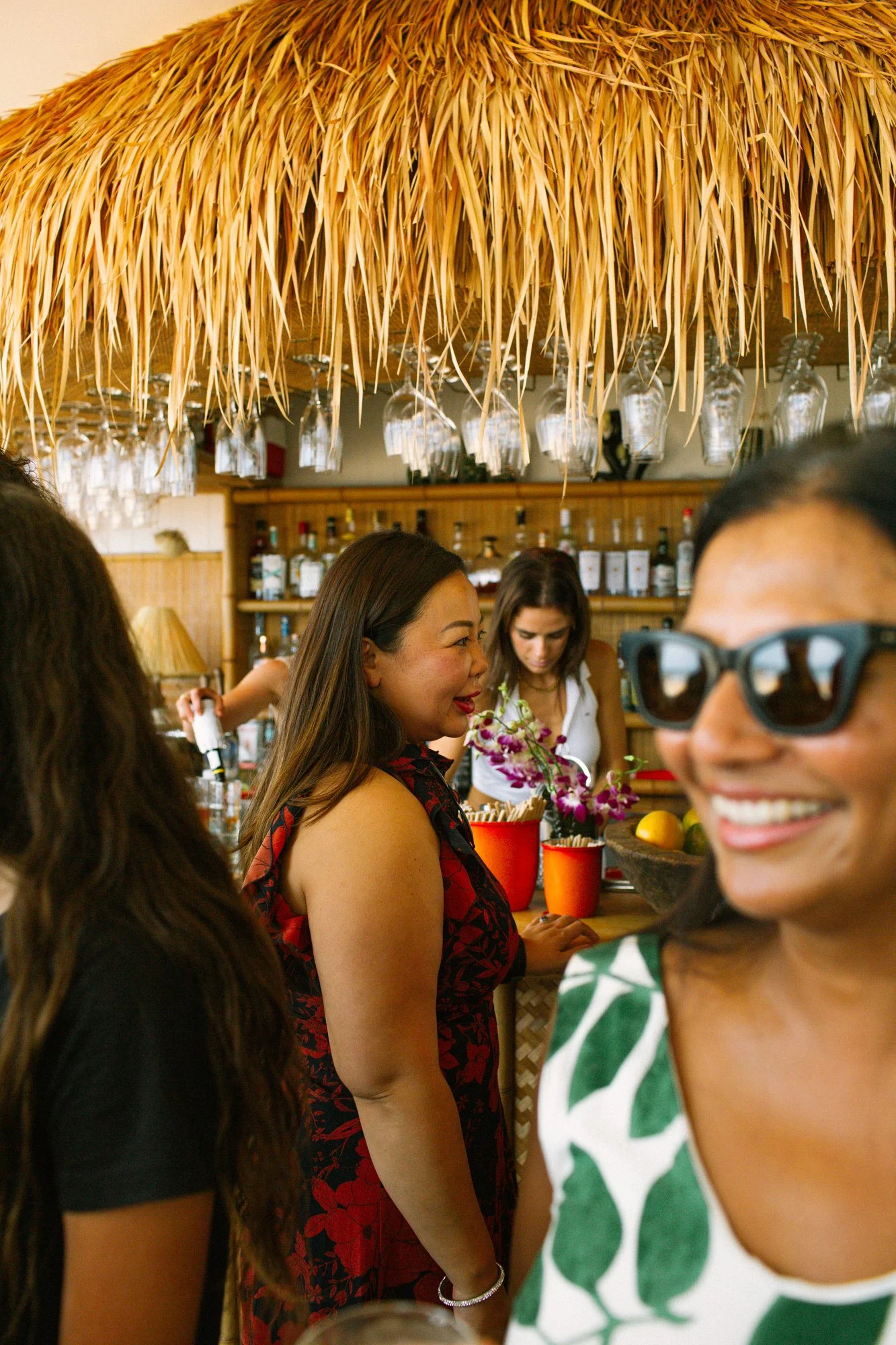 Group of women socializing at a tropical-themed bar with a thatched roof overhead.