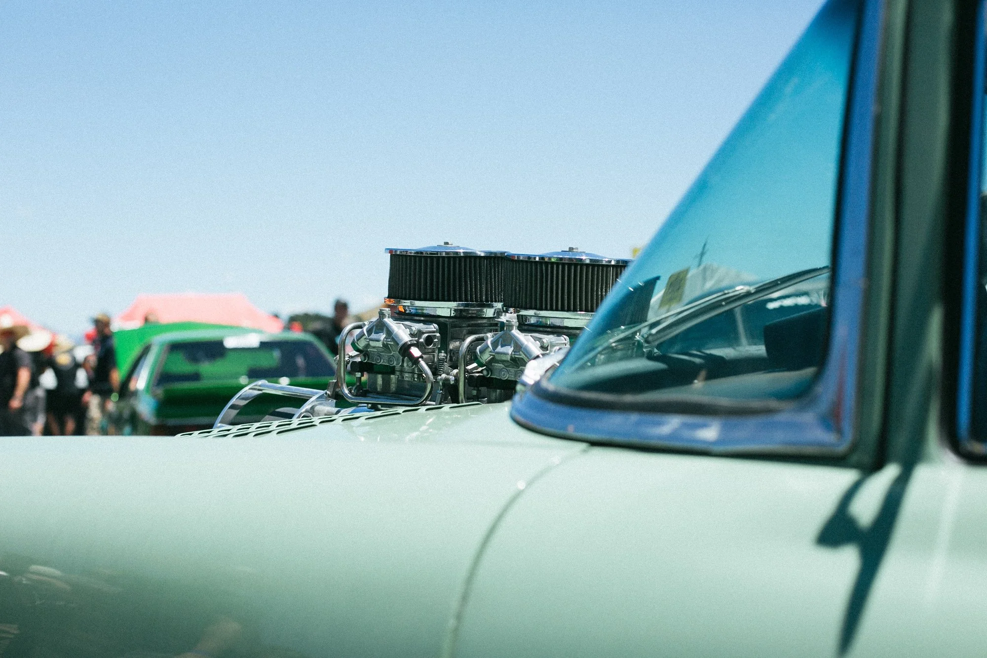 Close-up of a vintage car with an exposed engine and a large air intake, parked at an outdoor car event with people and tents in the background.