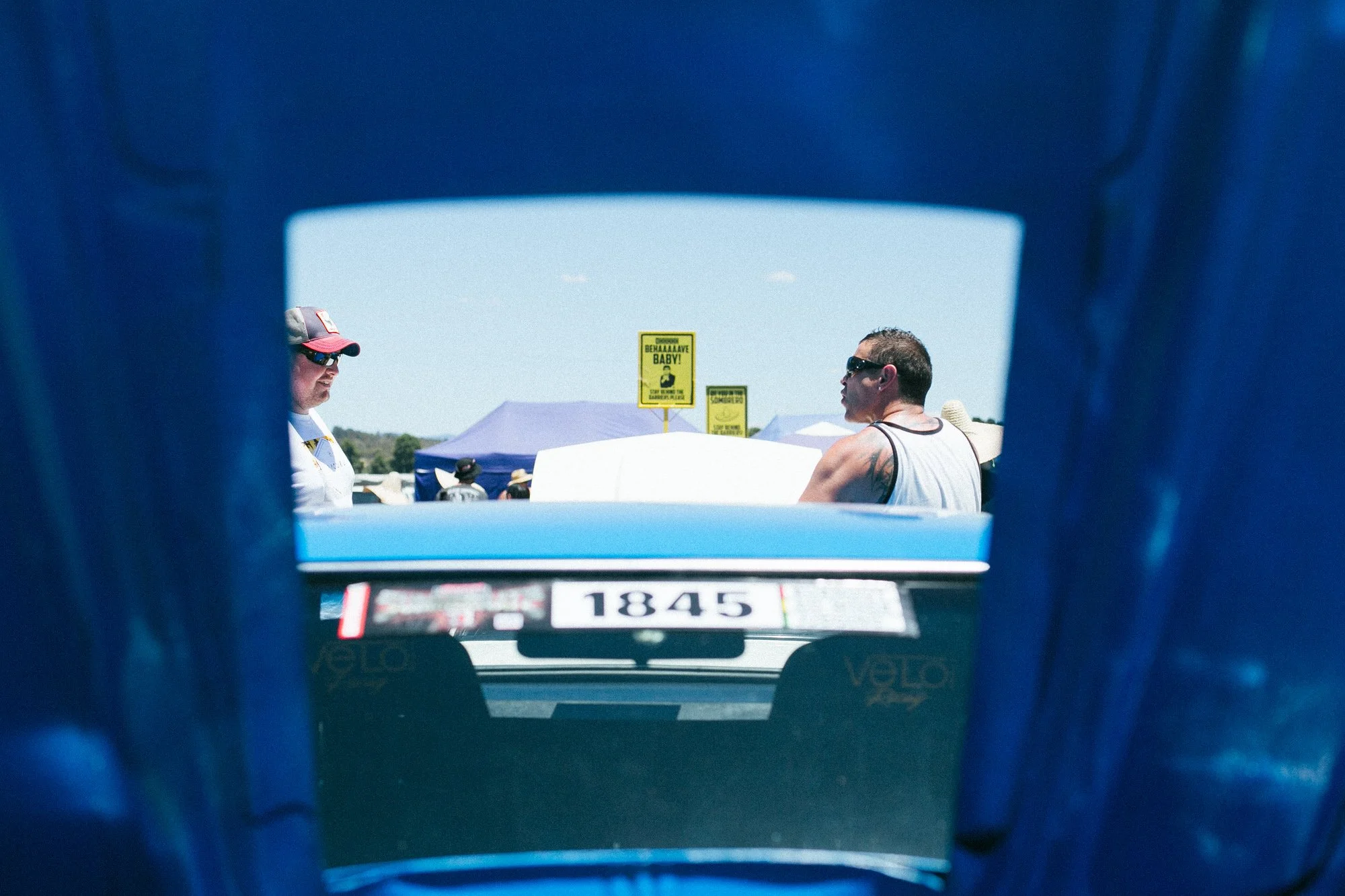 People at a racing event, viewed from behind a vehicle or barrier, with tents and signs in the background on a sunny day.