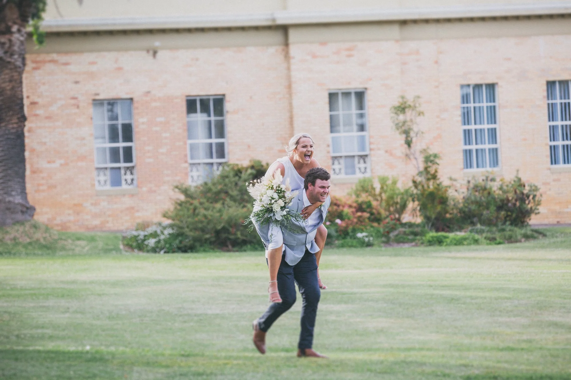 A happy couple enjoying a piggyback ride in a garden, with the woman holding a bouquet of flowers and laughing.
