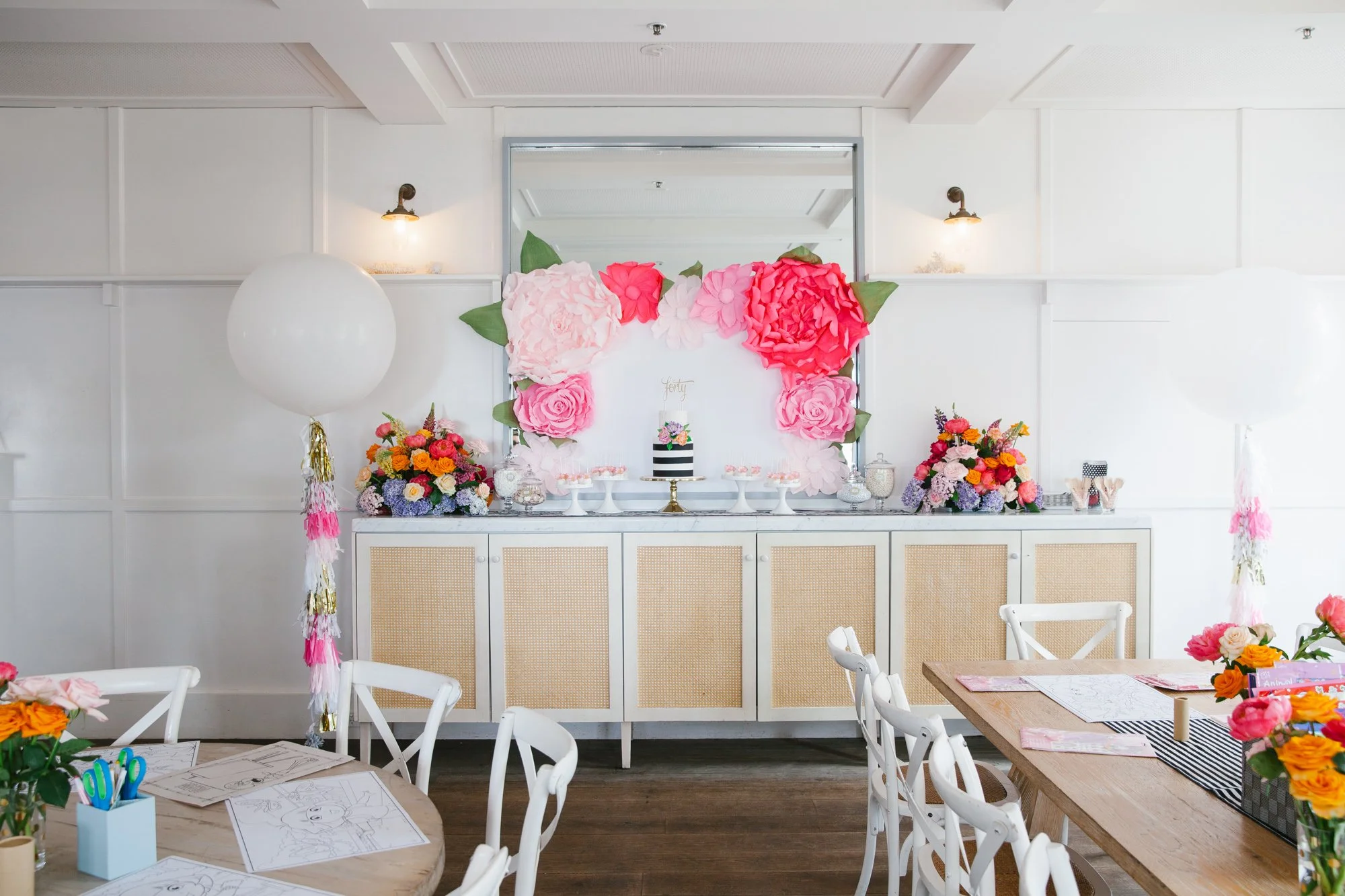 Decorative dessert table with pink and red paper flowers arch and floral arrangements, balloons, and party decorations in a bright room with wooden tables and white chairs.