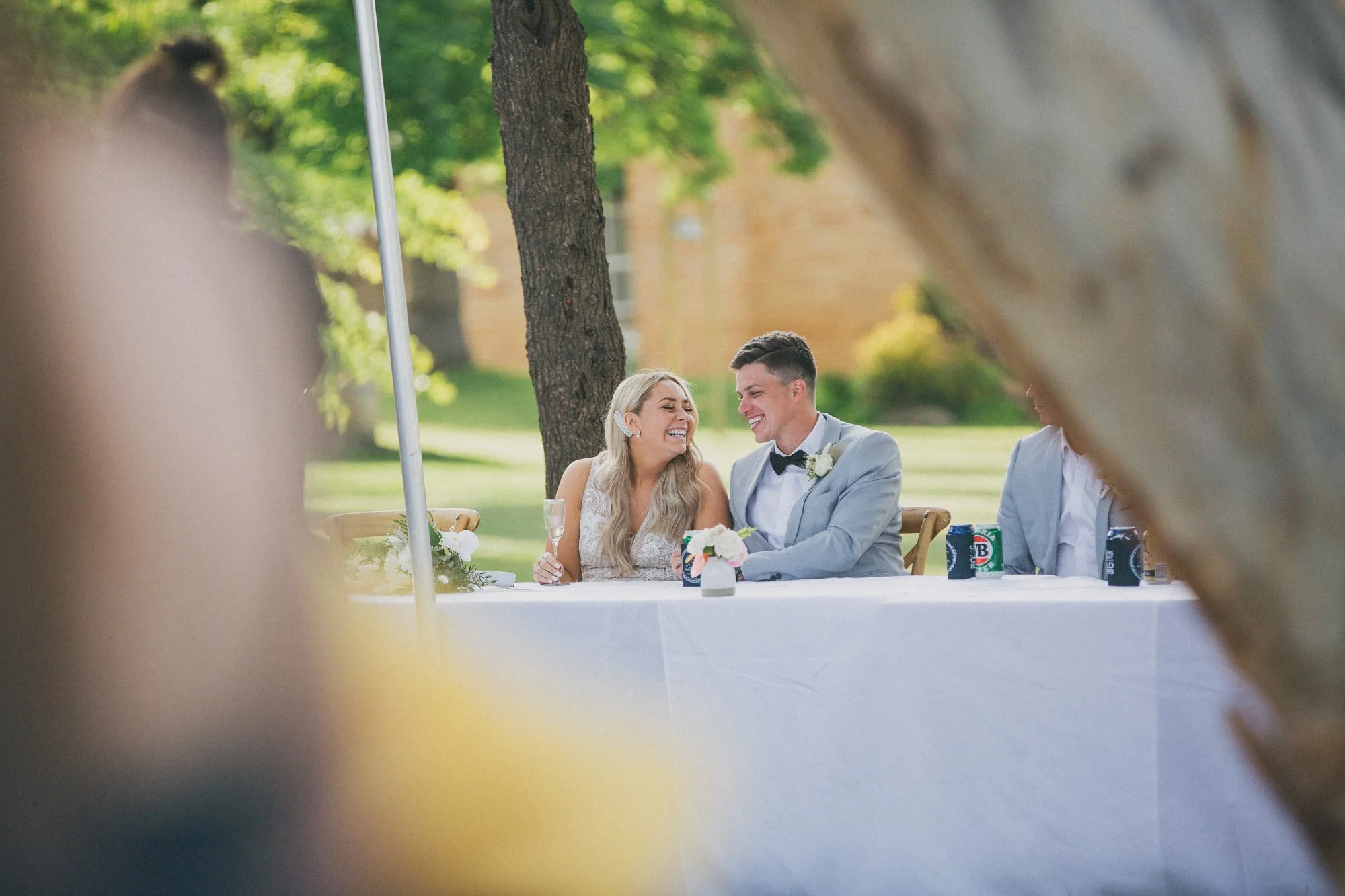 A bride and groom sitting at a wedding reception table outdoors, smiling and laughing, with glasses of champagne, a small flower arrangement, and drinks on the table. The scene is framed by tree trunks and a grassy background.