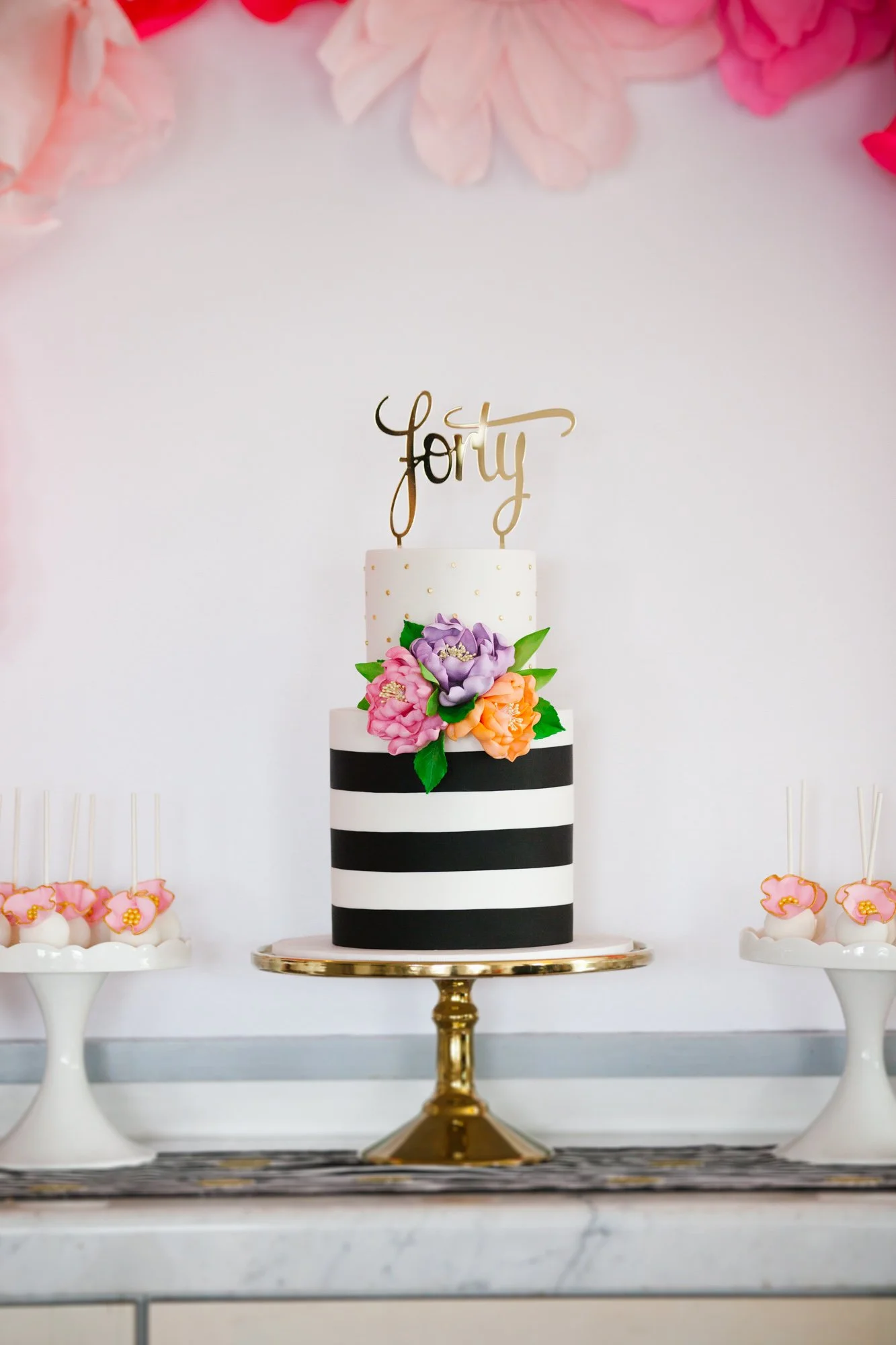 Elegant black and white striped birthday cake with purple, pink, and orange flowers, gold "forty" cake topper, displayed on a gold cake stand, with pink floral decorations above.
