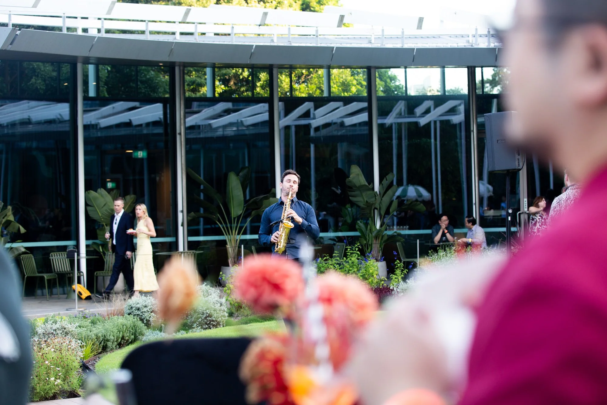 A live outdoor jazz performance featuring a saxophonist in a suit, with an audience and people mingling near a modern glass building with lush greenery.