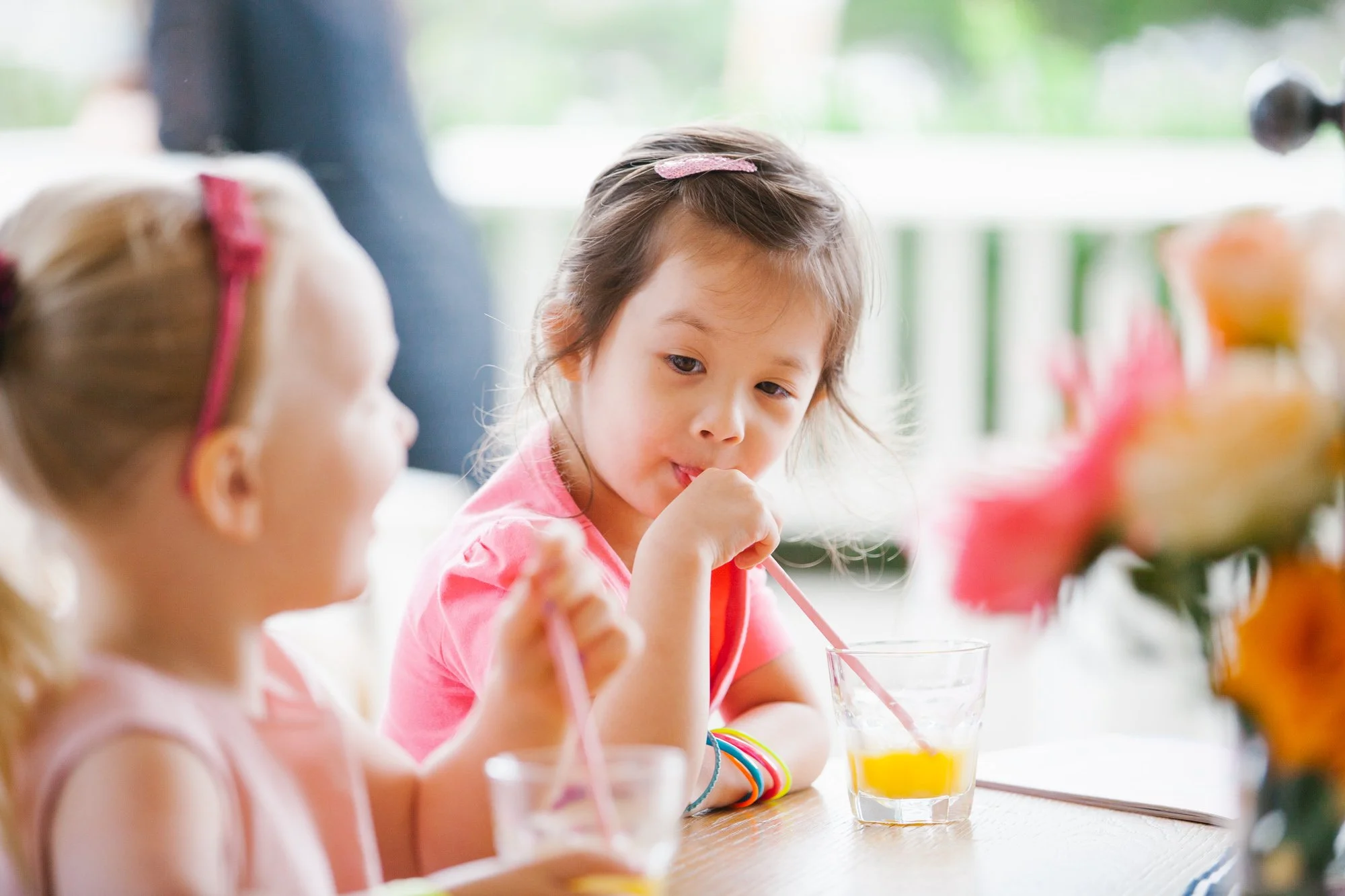 Two young girls sitting at a table, drinking juice from glasses with pink straws, with a blurred background of a green outdoor setting and a rose bouquet in the foreground.