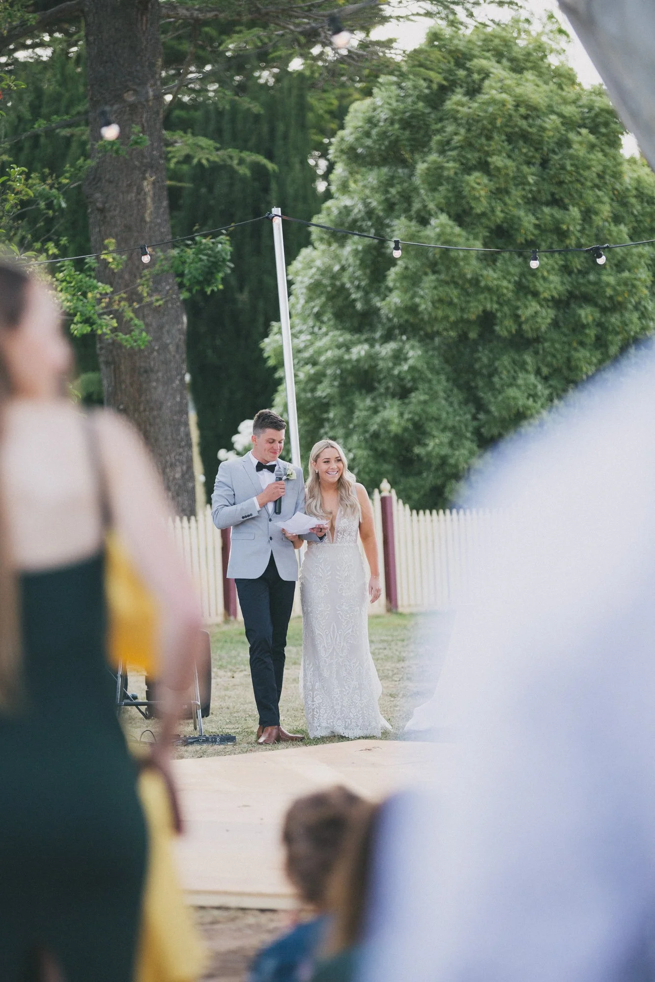A couple getting married outdoors, with the groom reading vows and the bride smiling. Guests are visible in the foreground, with green trees and string lights in the background.