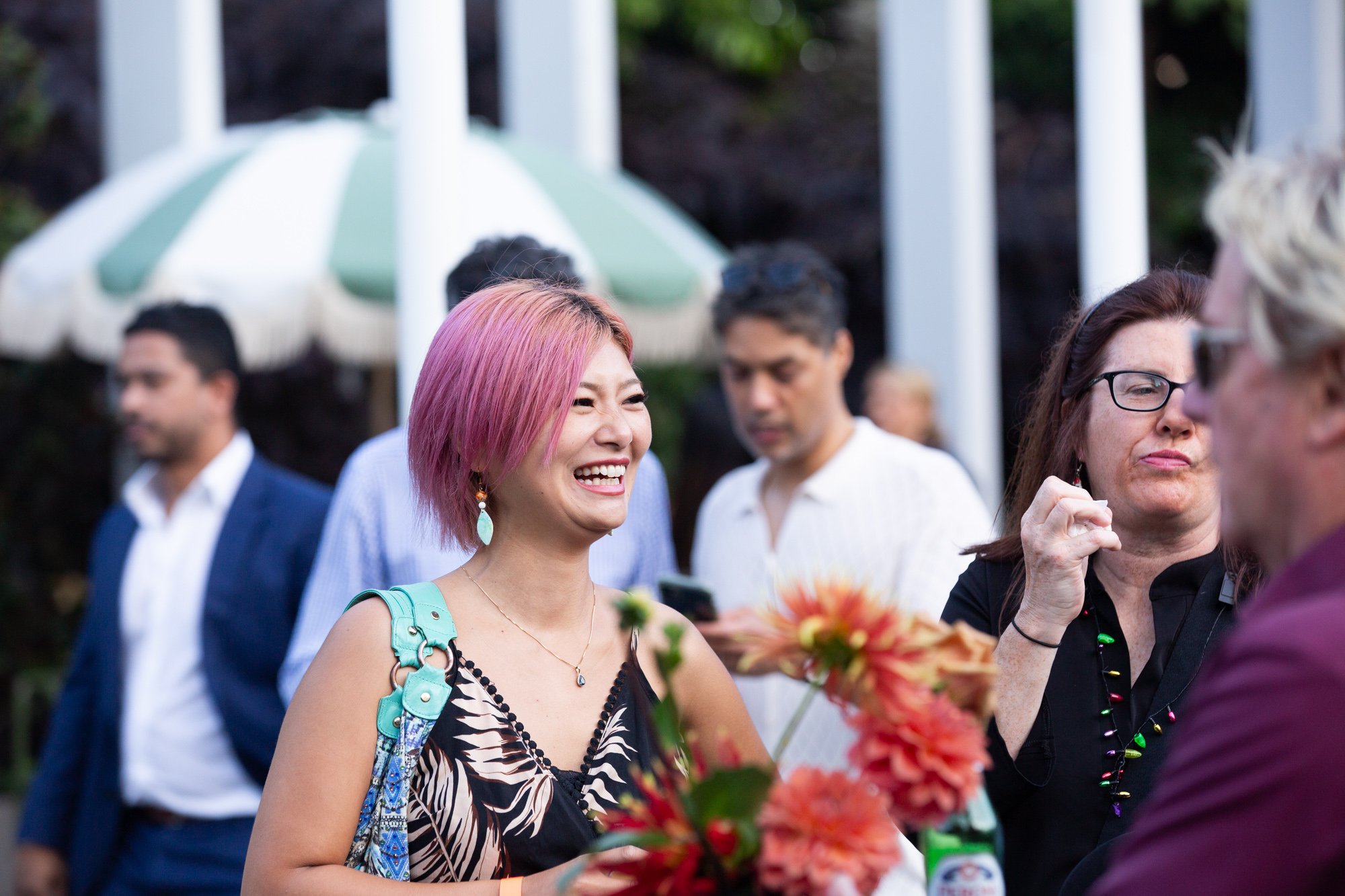 A group of people at an outdoor gathering, with a woman with pink hair smiling and talking to another person. Others are in the background near a green and white umbrella.