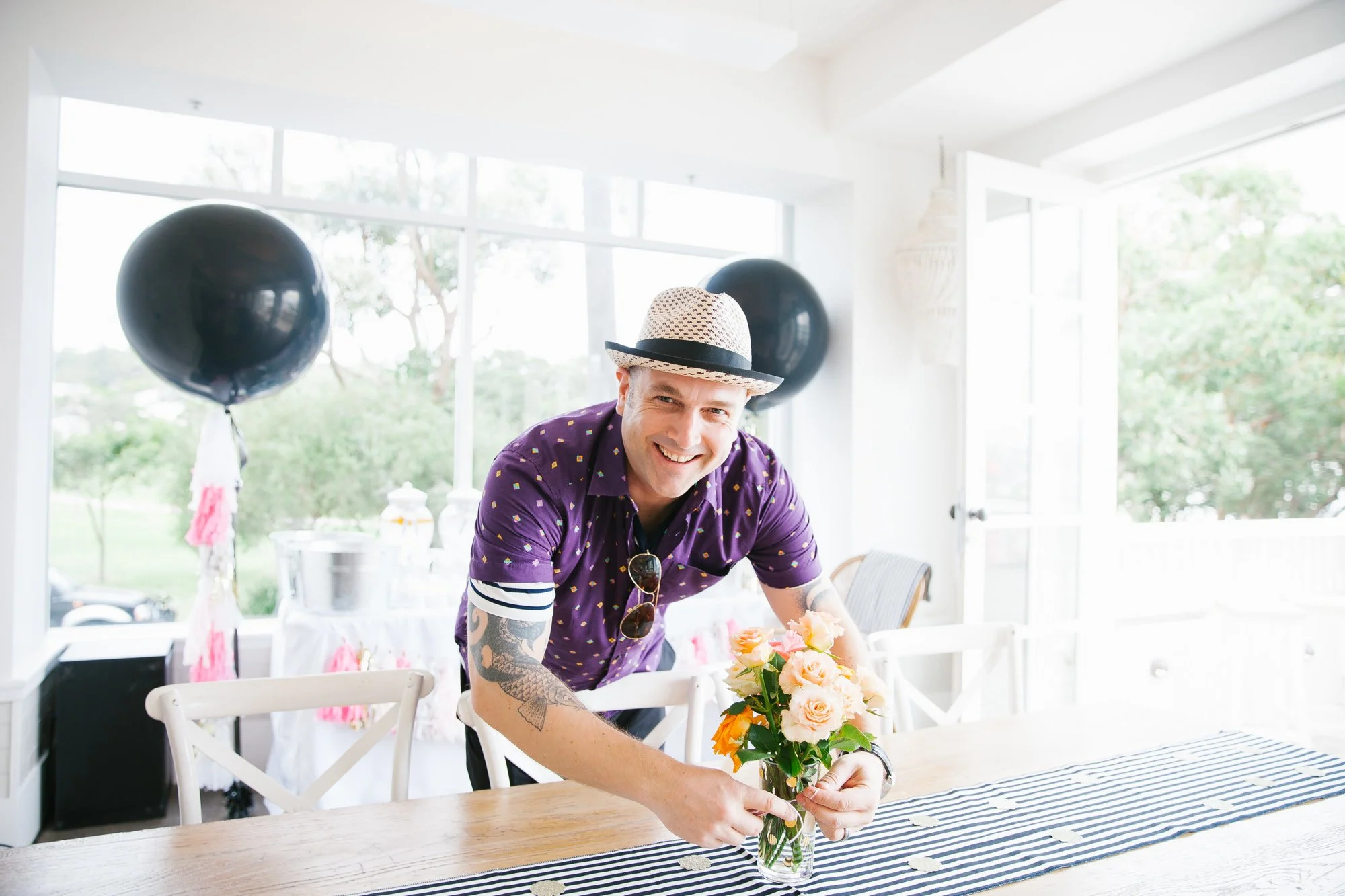 A smiling man in a purple shirt with yellow patterns and a straw hat, holding a bouquet of pink and orange flowers in a glass vase, in a bright room with large windows and black balloons in the background.