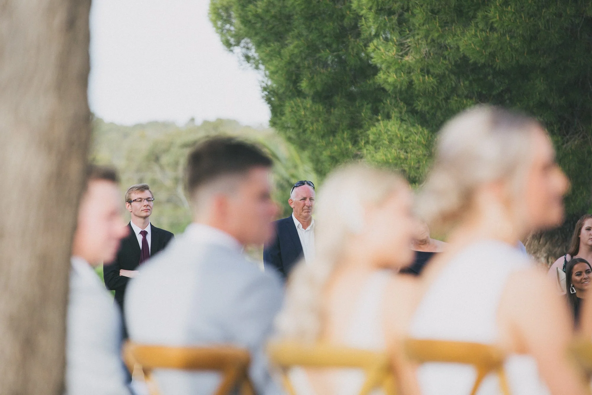 People attending an outdoor event, with some blurred individuals in the foreground and a man in a suit and sunglasses in the background, among trees and green foliage.