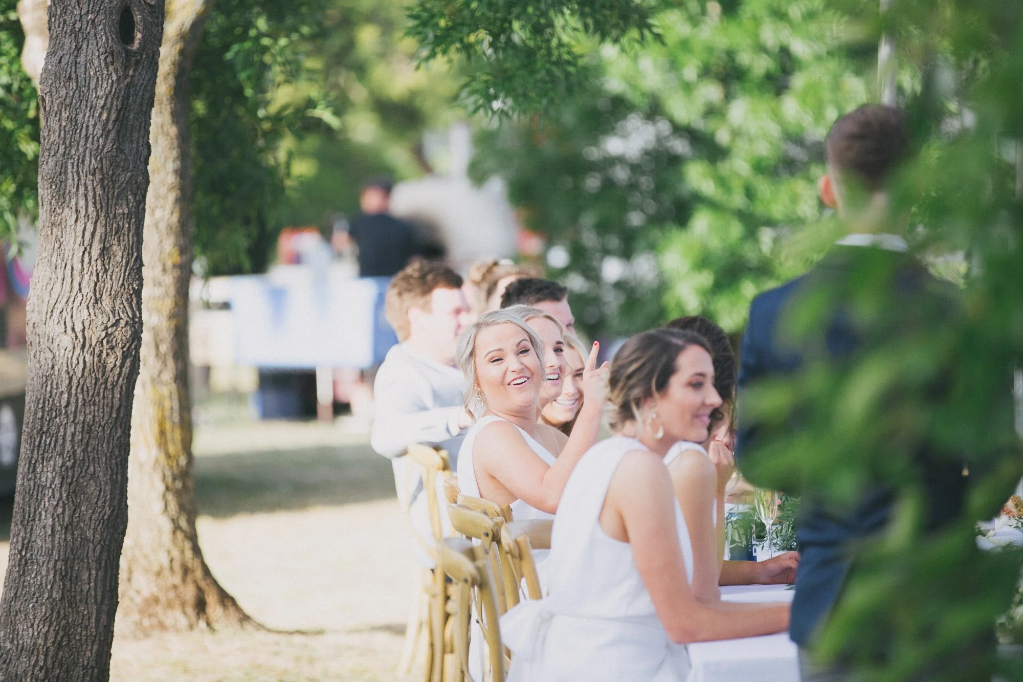 People sitting at a long outdoor table during a celebration or wedding, with trees and greenery in the background.