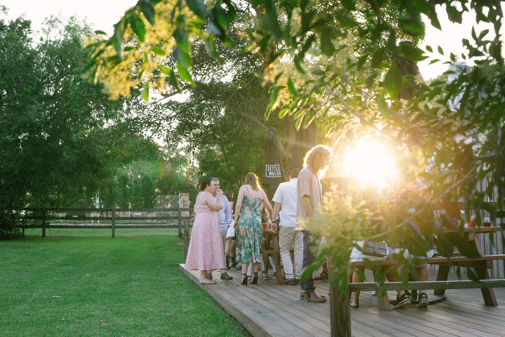 A group of people gathered on a wooden deck outdoors during sunset, surrounded by green trees and grass, with some sitting and others standing; the setting appears to be a social event.