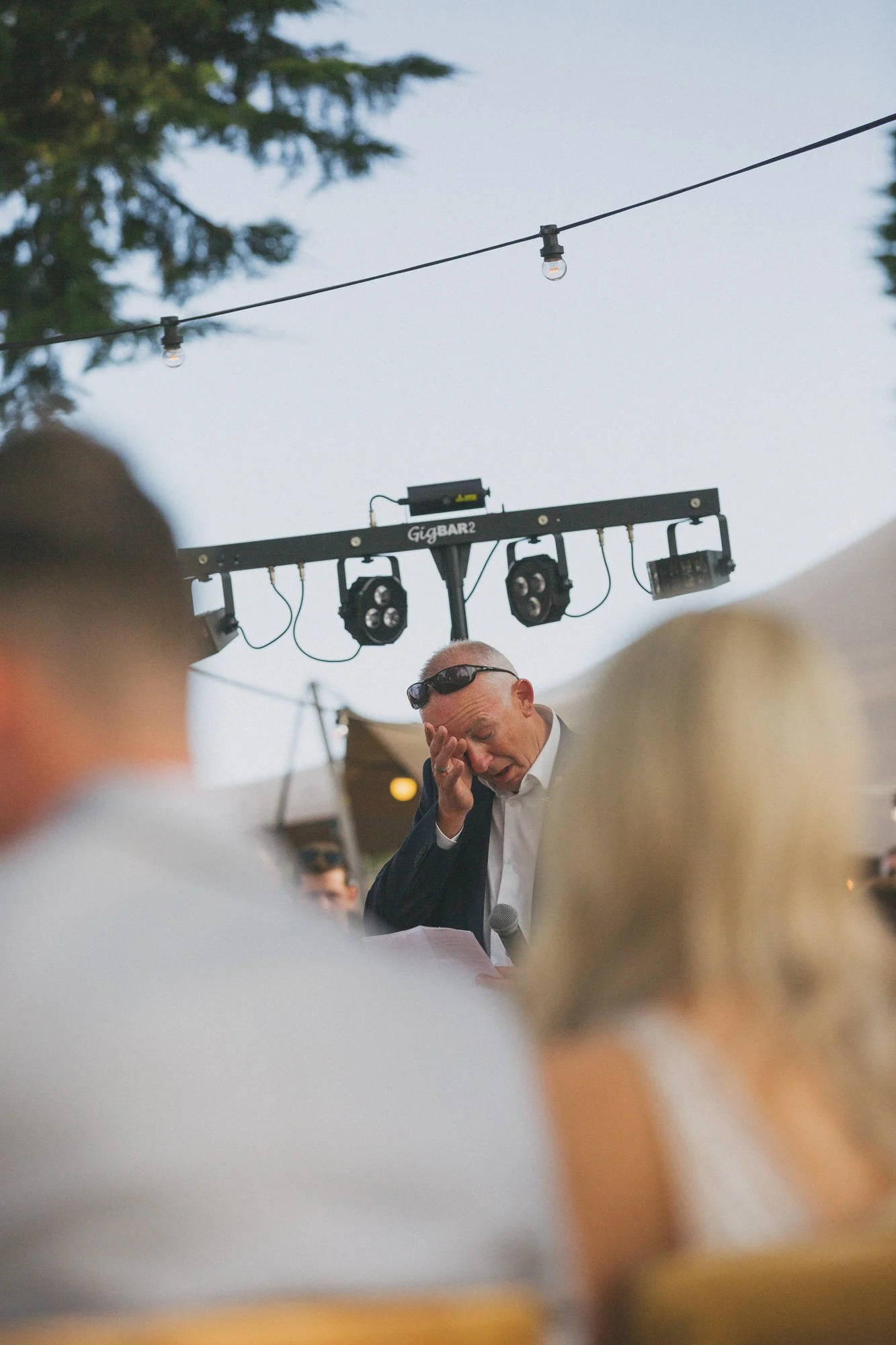 A man with sunglasses appears emotional or overwhelmed at an outdoor event, standing in front of a microphone with a crowd in the foreground and stage lighting above.