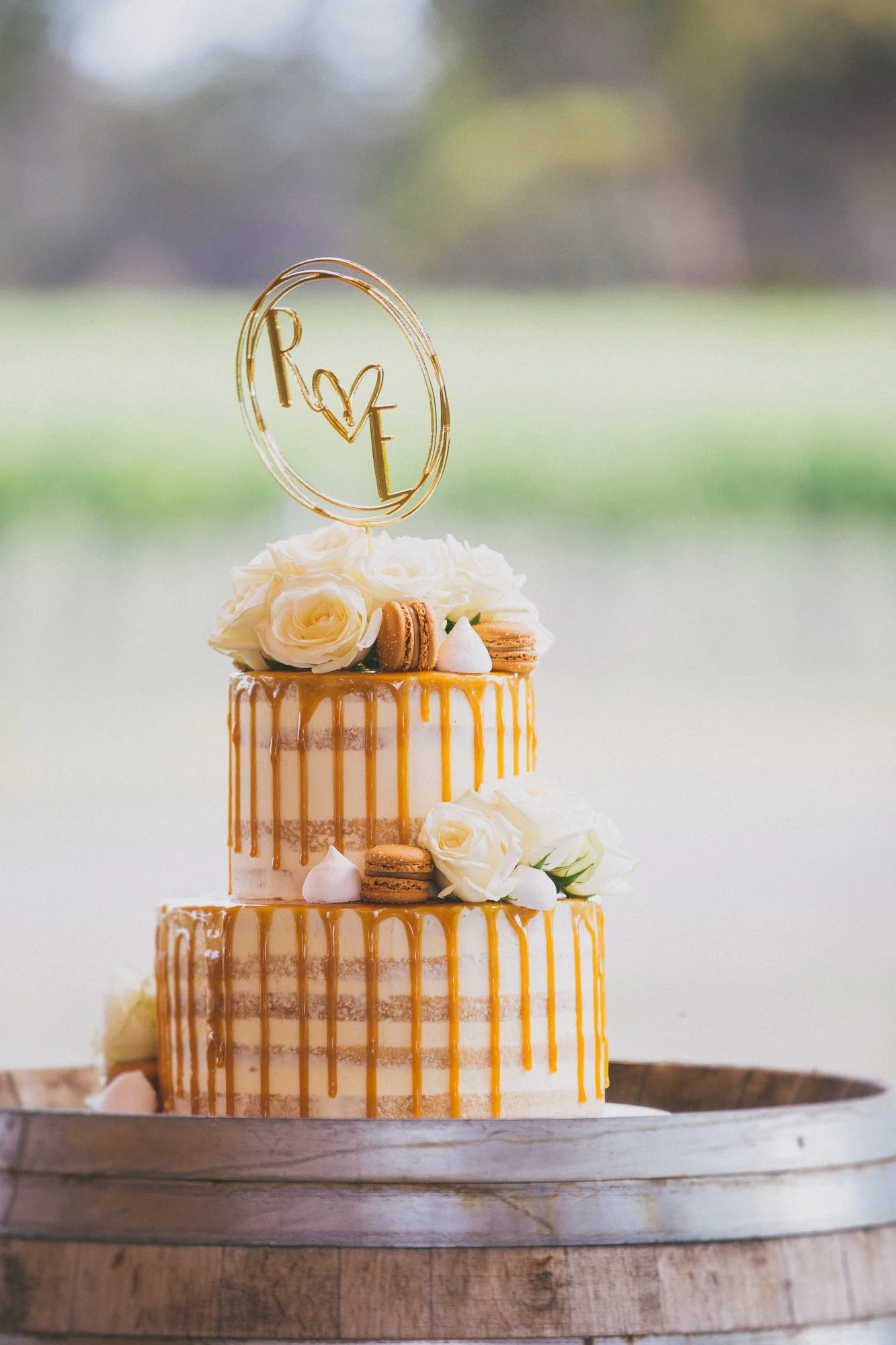 A two-tiered wedding cake decorated with white roses, caramel macarons, small white meringues, and gold drip icing, topped with a custom cake topper spelling "LOVE" with a heart, on a wooden stand with a blurred outdoor background.