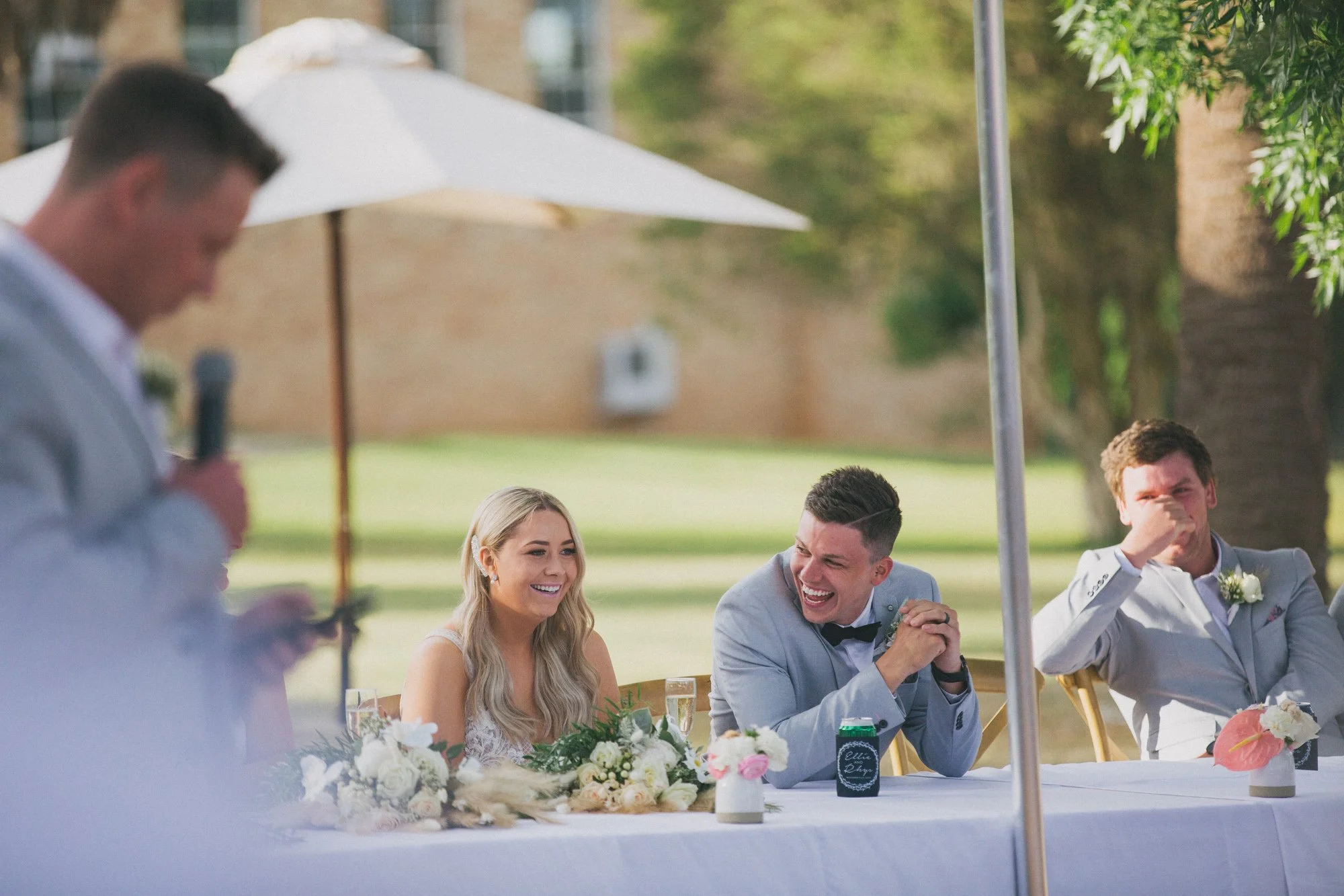 A wedding reception outdoors with three people sitting at a decorated table: a blonde woman in a white dress, a man in a light gray suit with a bow tie, and another man in a similar suit. An older man is speaking at a microphone in the foreground. Th