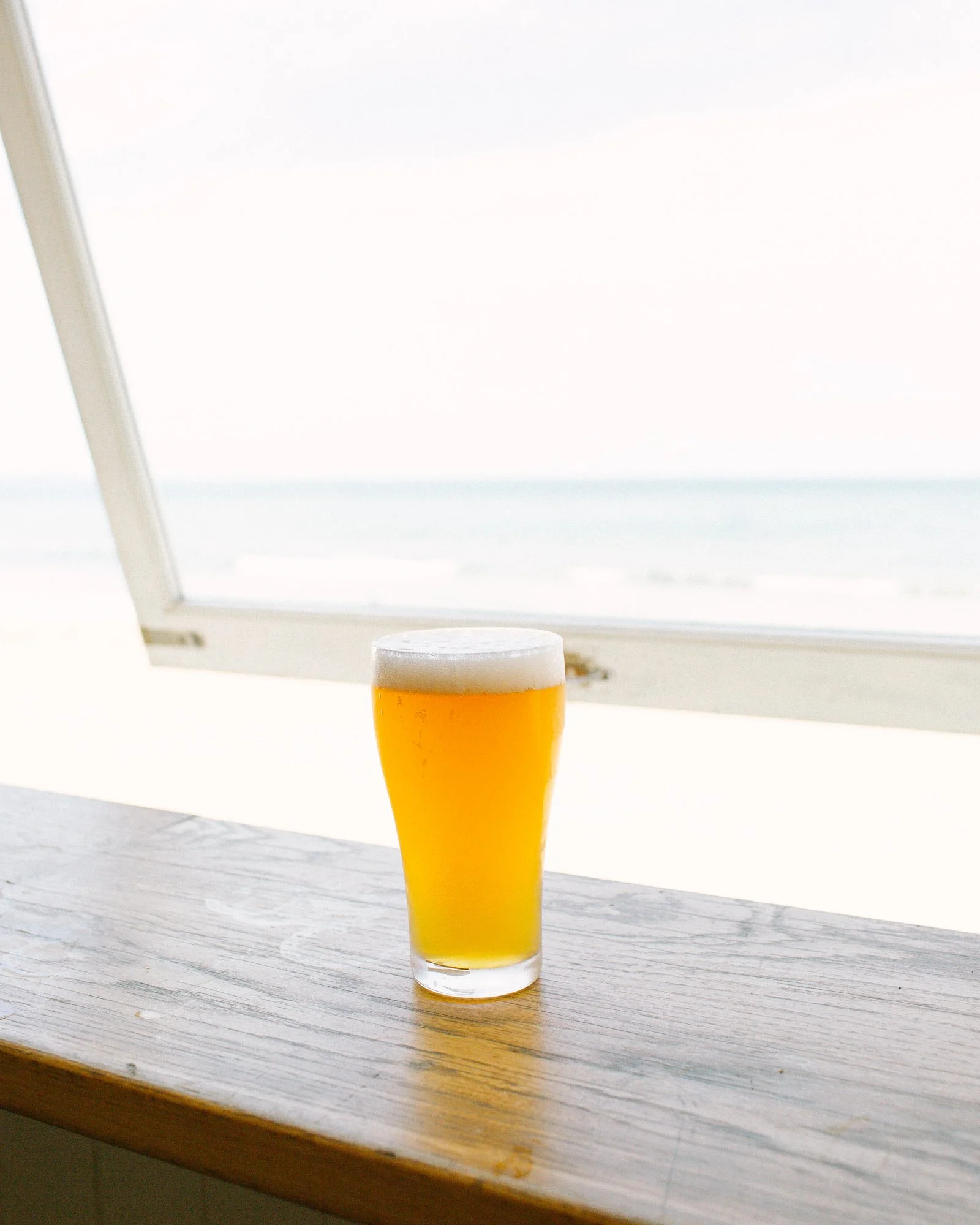 A glass of beer on a wooden windowsill with a beach view in the background.