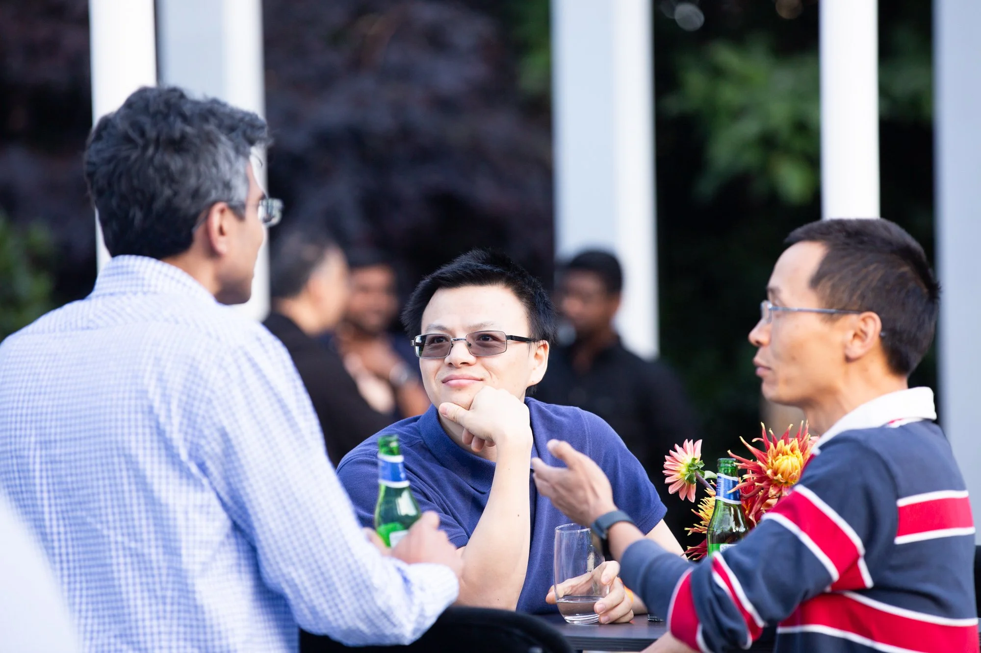 Three men engaged in conversation at an outdoor event, with one smiling and resting his chin on his hand, drinking a beverage, and the others holding drinks, surrounded by other people and flowers in the background.
