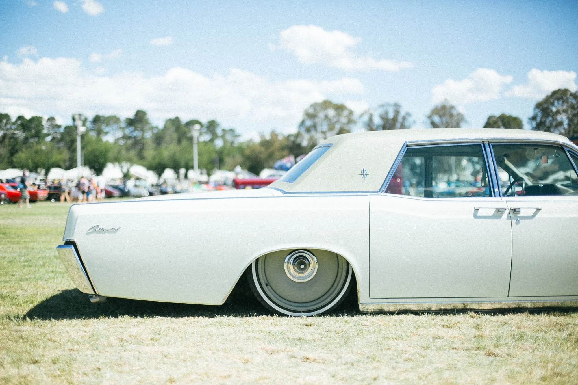 A vintage white car with a lowered body and custom wheels parked on a grassy field during a car show, with other cars and people in the background under a blue sky with clouds.