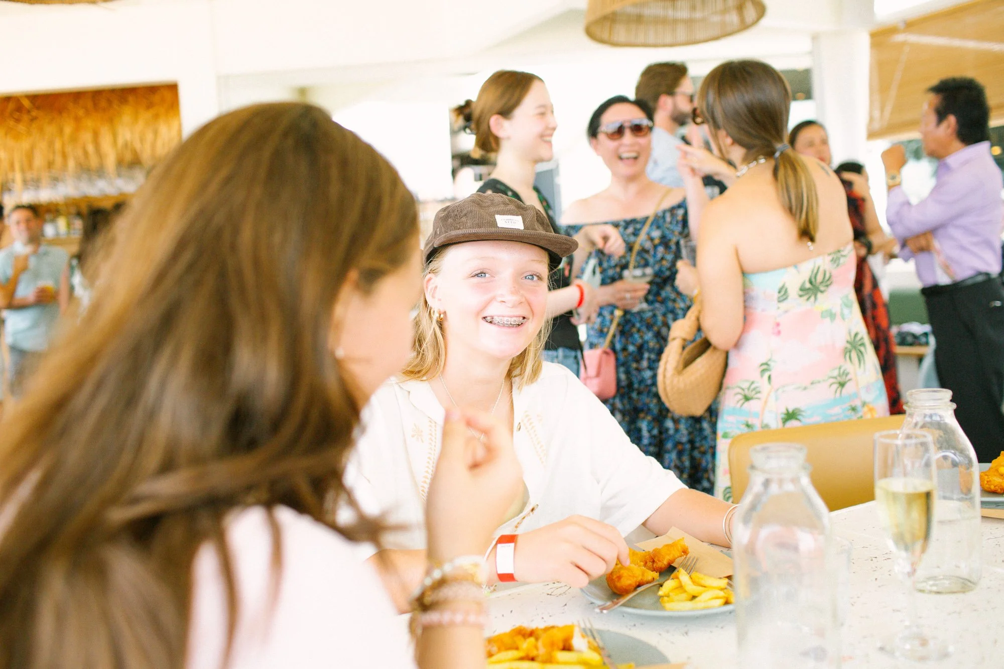 A girl with blond hair, braces, and a brown cap smiling at a lunch table with a plate of fried chicken and fries. In the background, a group of people are socializing, including women in summer dresses, some wearing sunglasses, and a man in a purple 