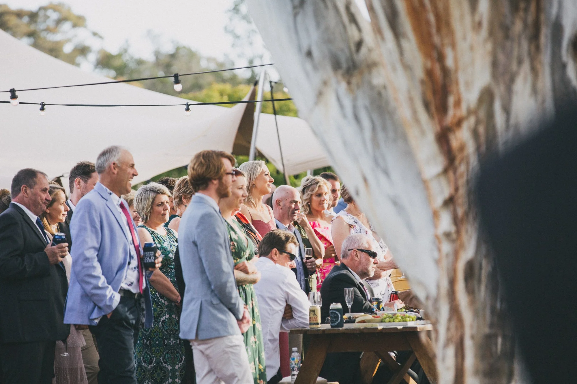 People attending an outdoor party or gathering, possibly a wedding, under a large white tent with string lights, with a wood table and some drinks, surrounded by trees.