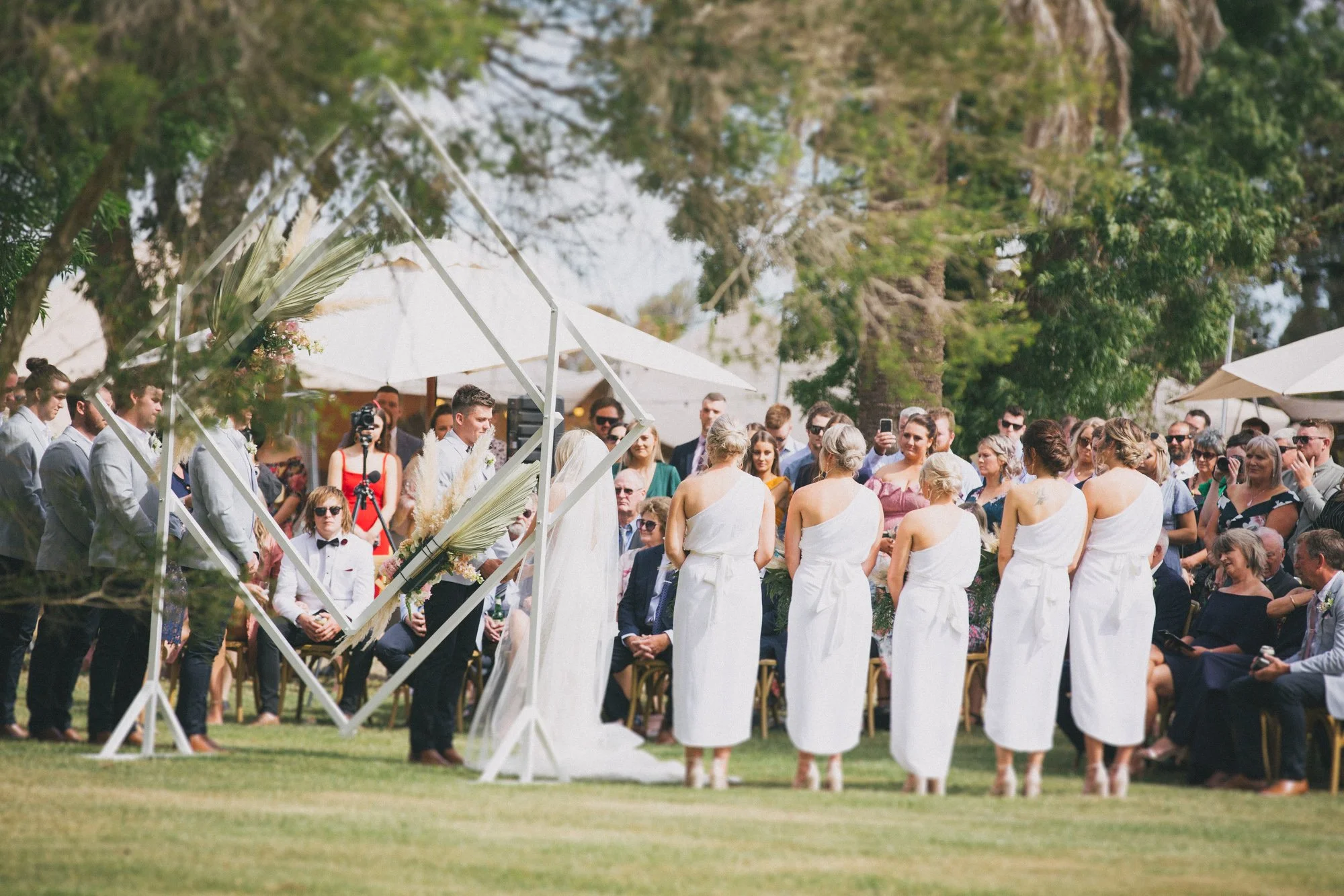 A wedding ceremony outdoors with five women dressed in white standing in front of a gathered crowd, facing the officiant. The setting is lush with trees and white umbrellas providing shade, and guests are seated and standing, many taking photos.