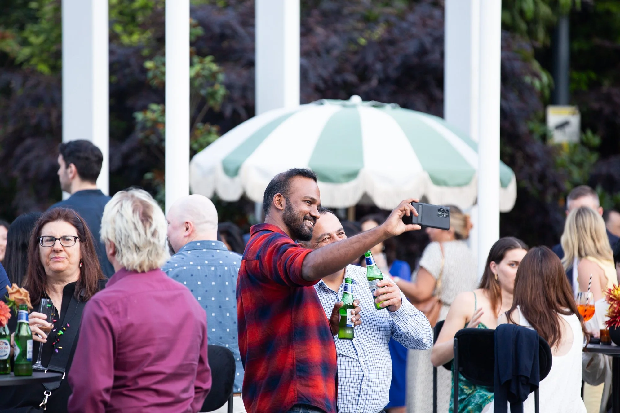 People at an outdoor social event, some holding drinks, with a man in a red plaid shirt taking a selfie in front of a green and white umbrella.