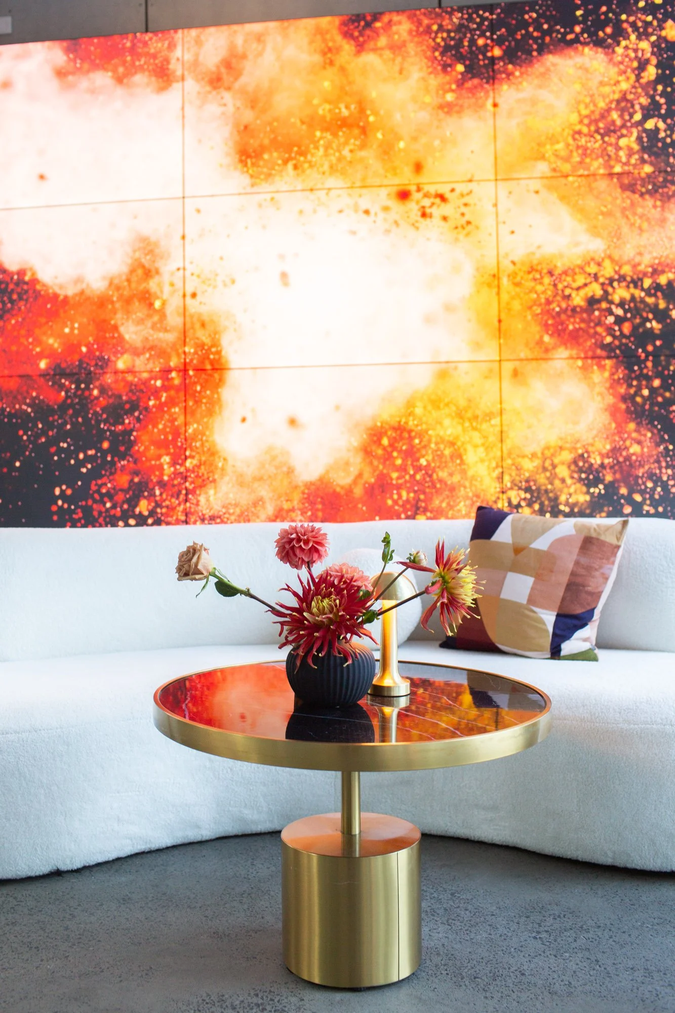 Modern living room with a white sofa, a round gold and black coffee table, a vase of pink and cream flowers, a gold lamp, and a colorful abstract wall art featuring fiery orange, red, yellow, and white hues.