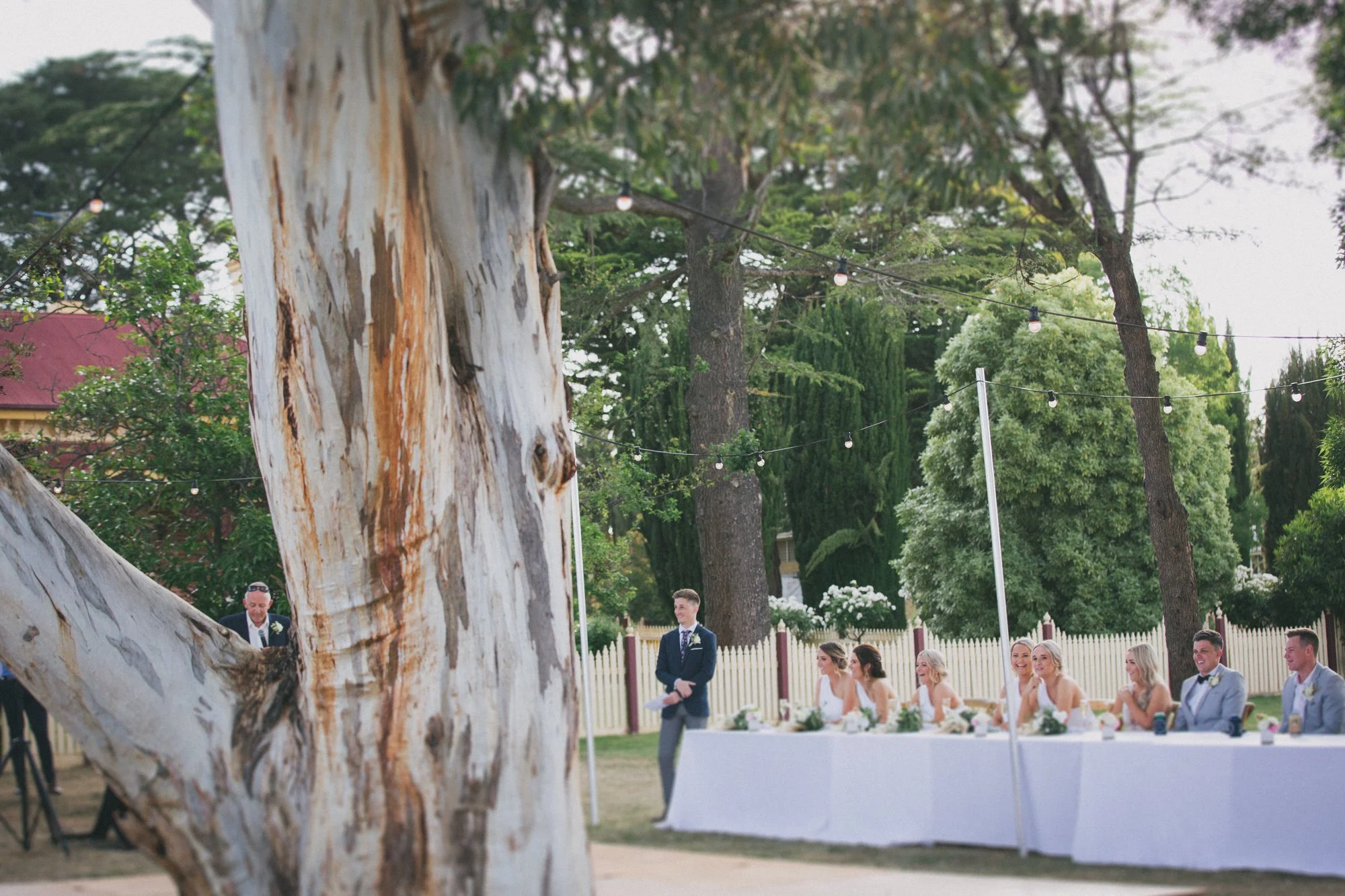 Outdoor wedding reception under string lights, with a long table of seated guests, some women wearing white dresses, and a man standing nearby.