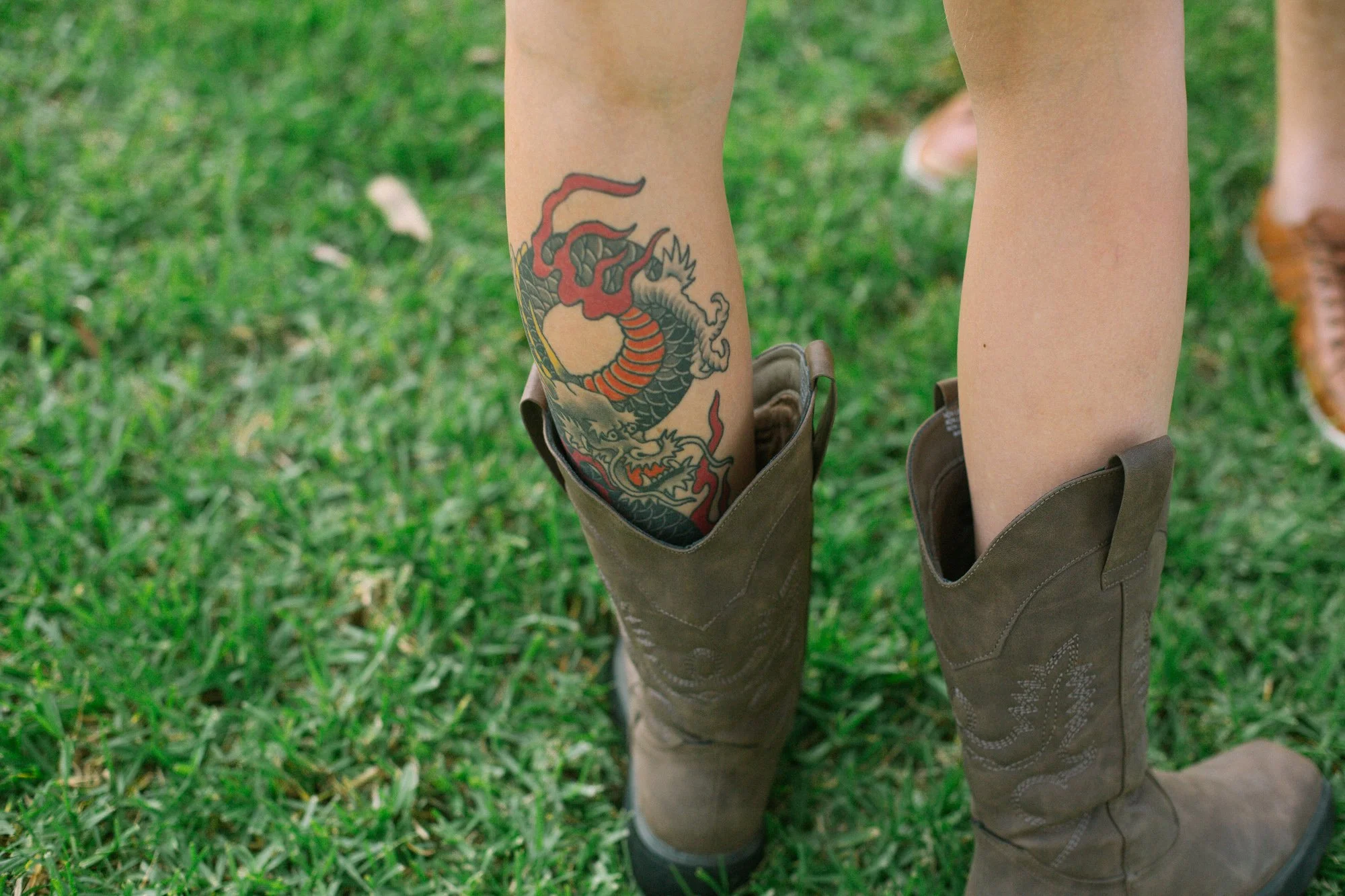 Person wearing cowboy boots with a colorful dragon tattoo on their calf, standing on a grassy field.