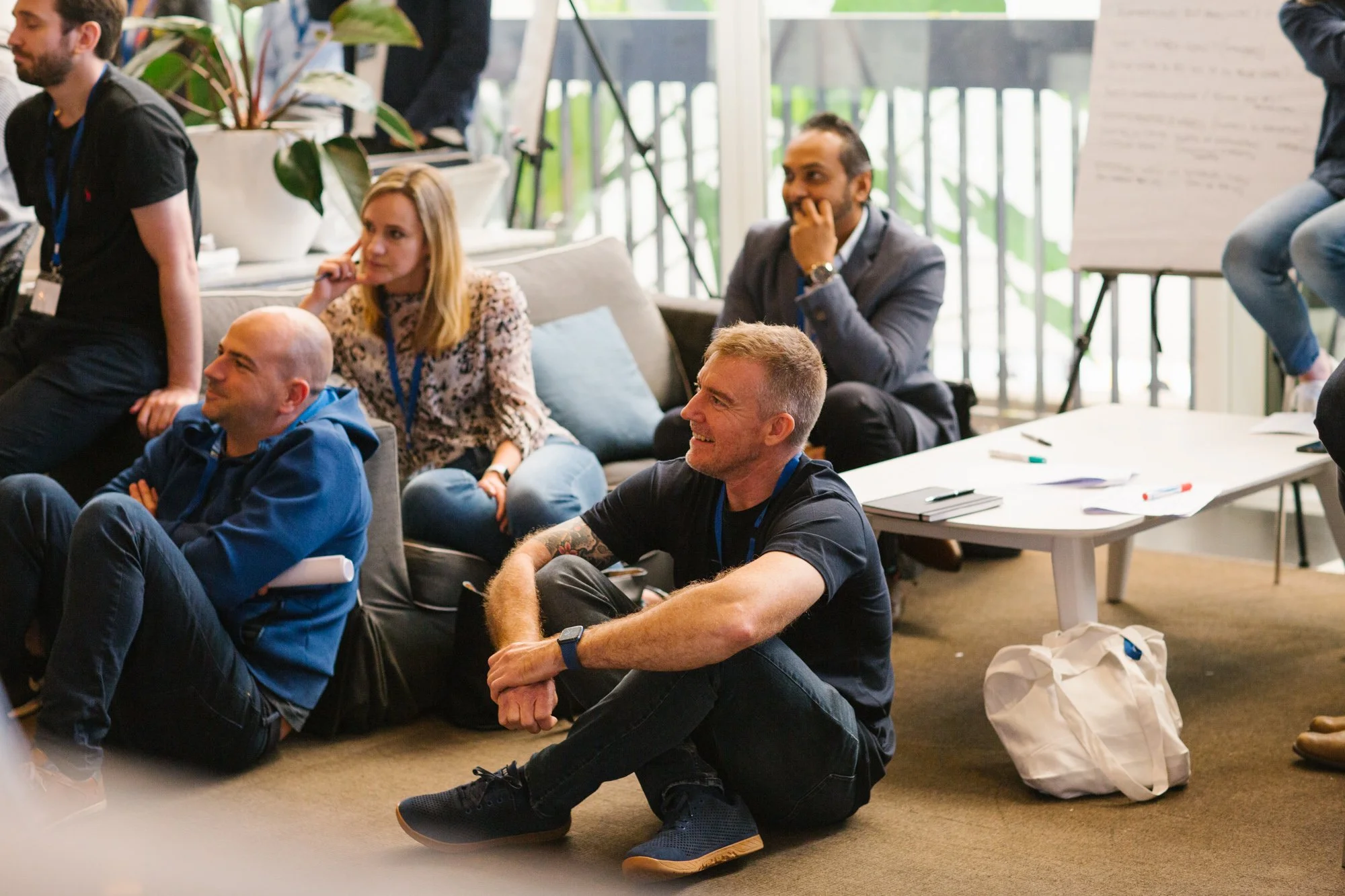 People sitting and listening during a presentation or meeting in a bright room with large windows, some seated on a sofa, others on the floor and a low table with notebooks and pens.