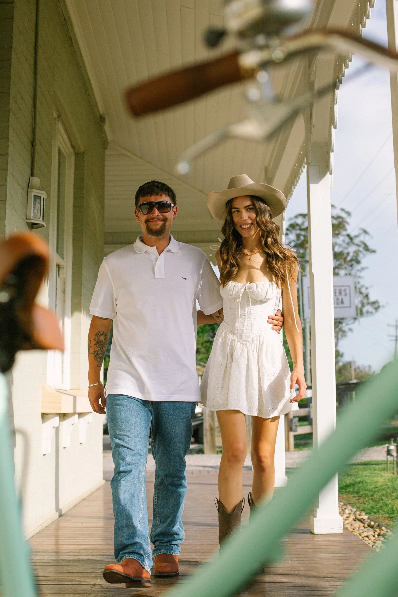 A smiling man and woman walking arm in arm on a porch, with the man wearing sunglasses, a white polo shirt, and jeans, and the woman wearing a wide-brimmed hat and a white dress.