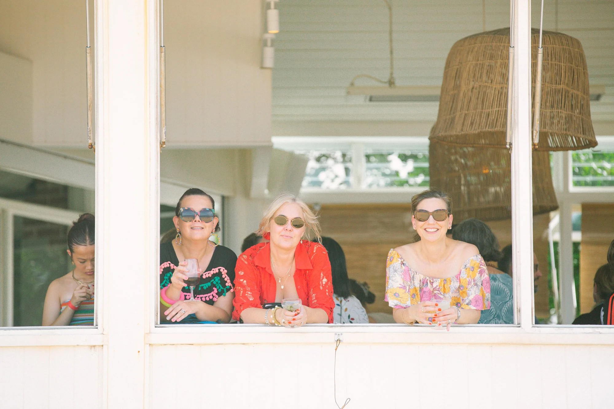 Three women wearing sunglasses at a window, holding drinks, smiling, with other people and a large hanging lamp visible inside.