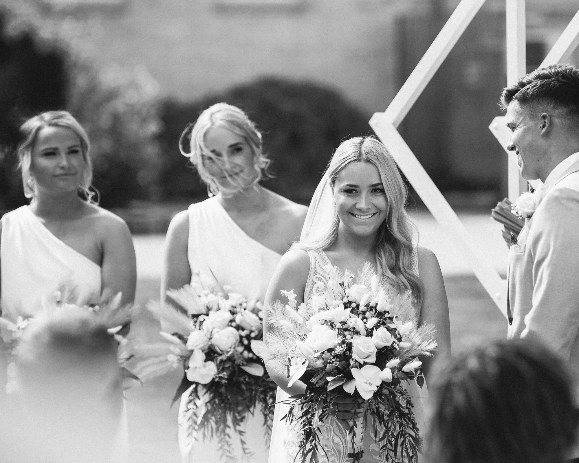 Black and white photo of a wedding ceremony with a smiling bride holding a large bouquet, standing next to the groom who is smiling at her. Two bridesmaids stand behind the bride, holding smaller bouquets, with the one on the left looking at the came
