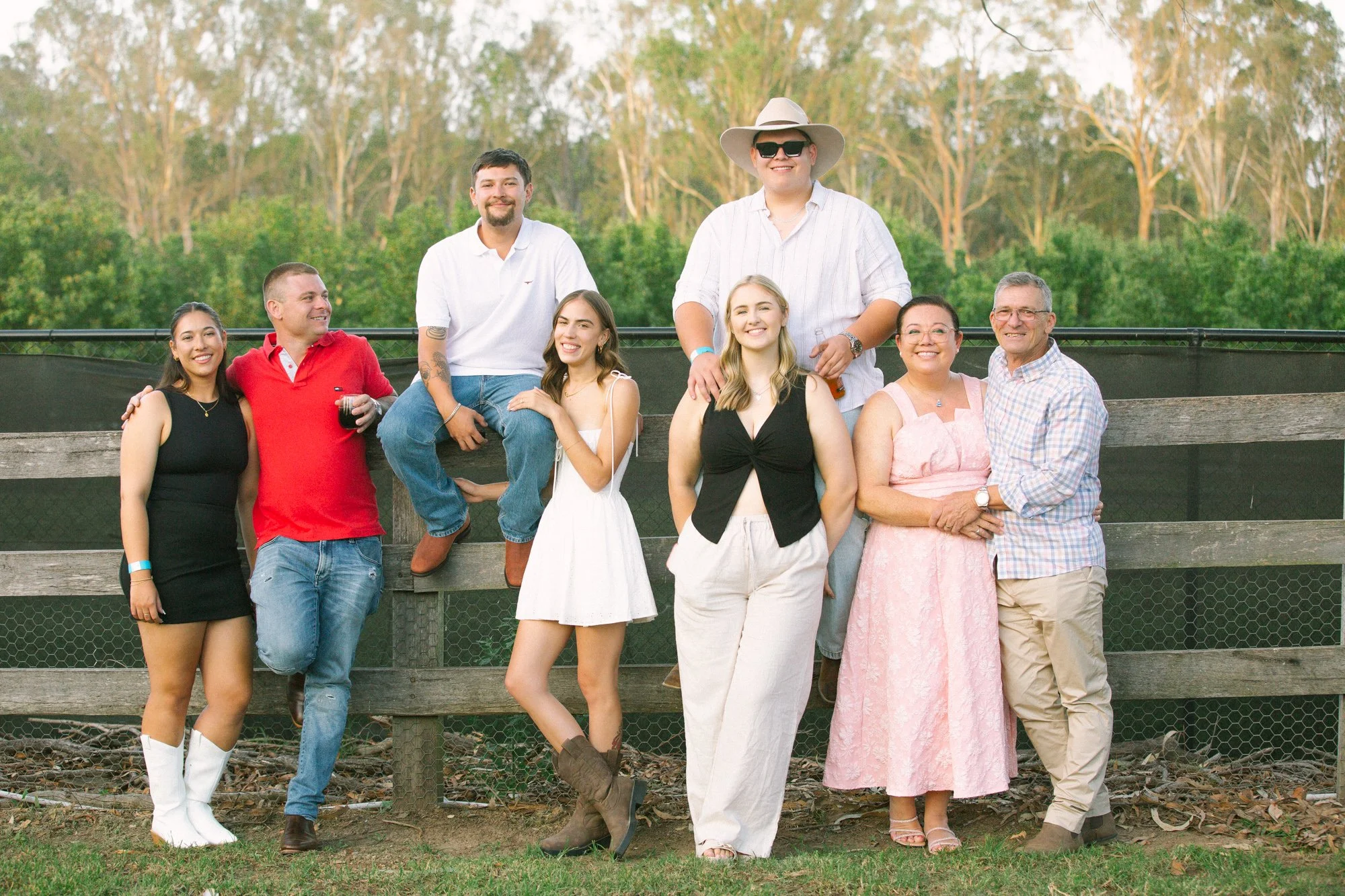 Group of nine people posing outdoors on a sunny day, with a wooden fence and trees in the background.