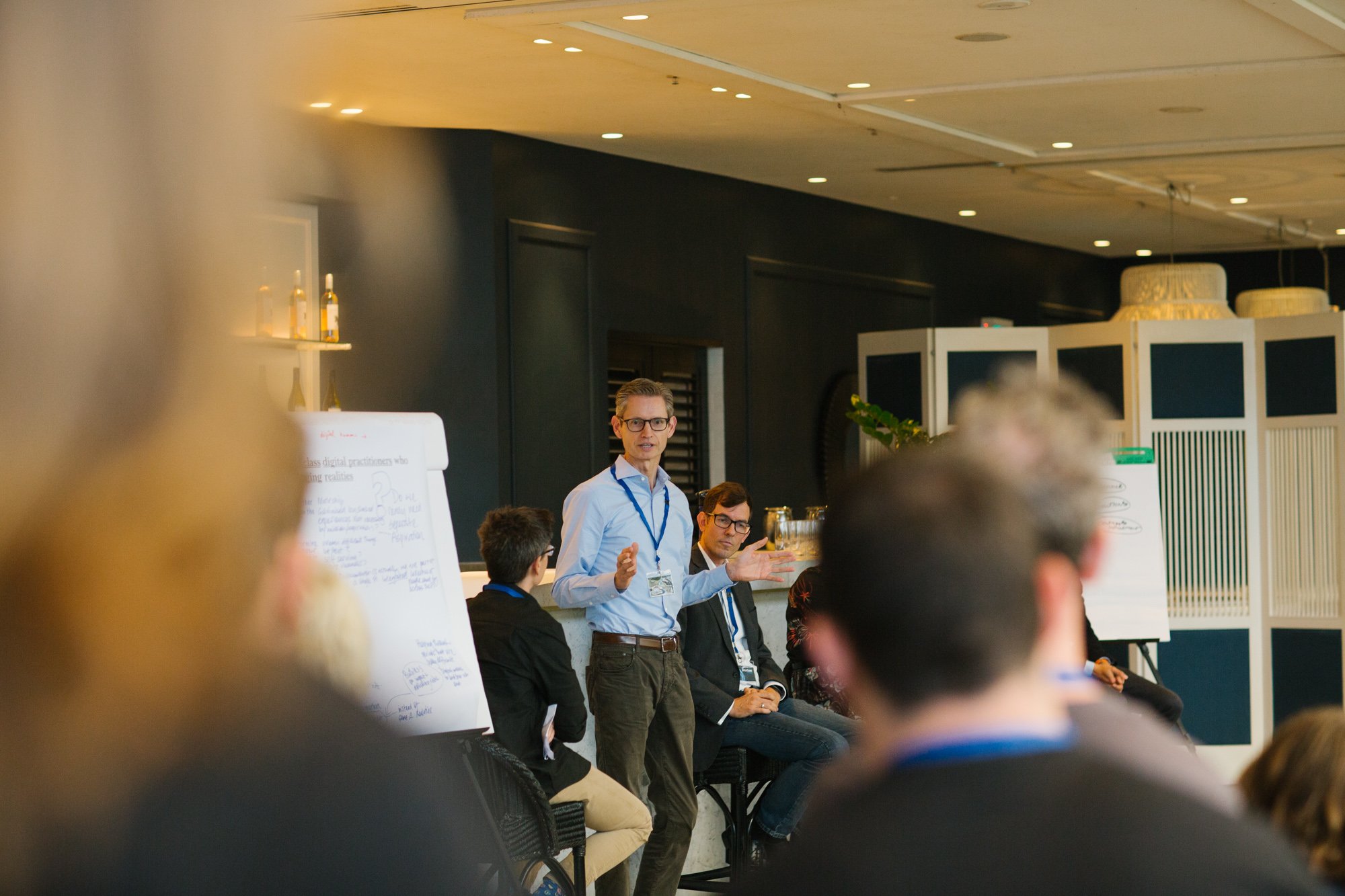 A man in glasses and a blue shirt speaking at a panel discussion or conference, with several other seated individuals, whiteboards, and a dark background in a well-lit room.