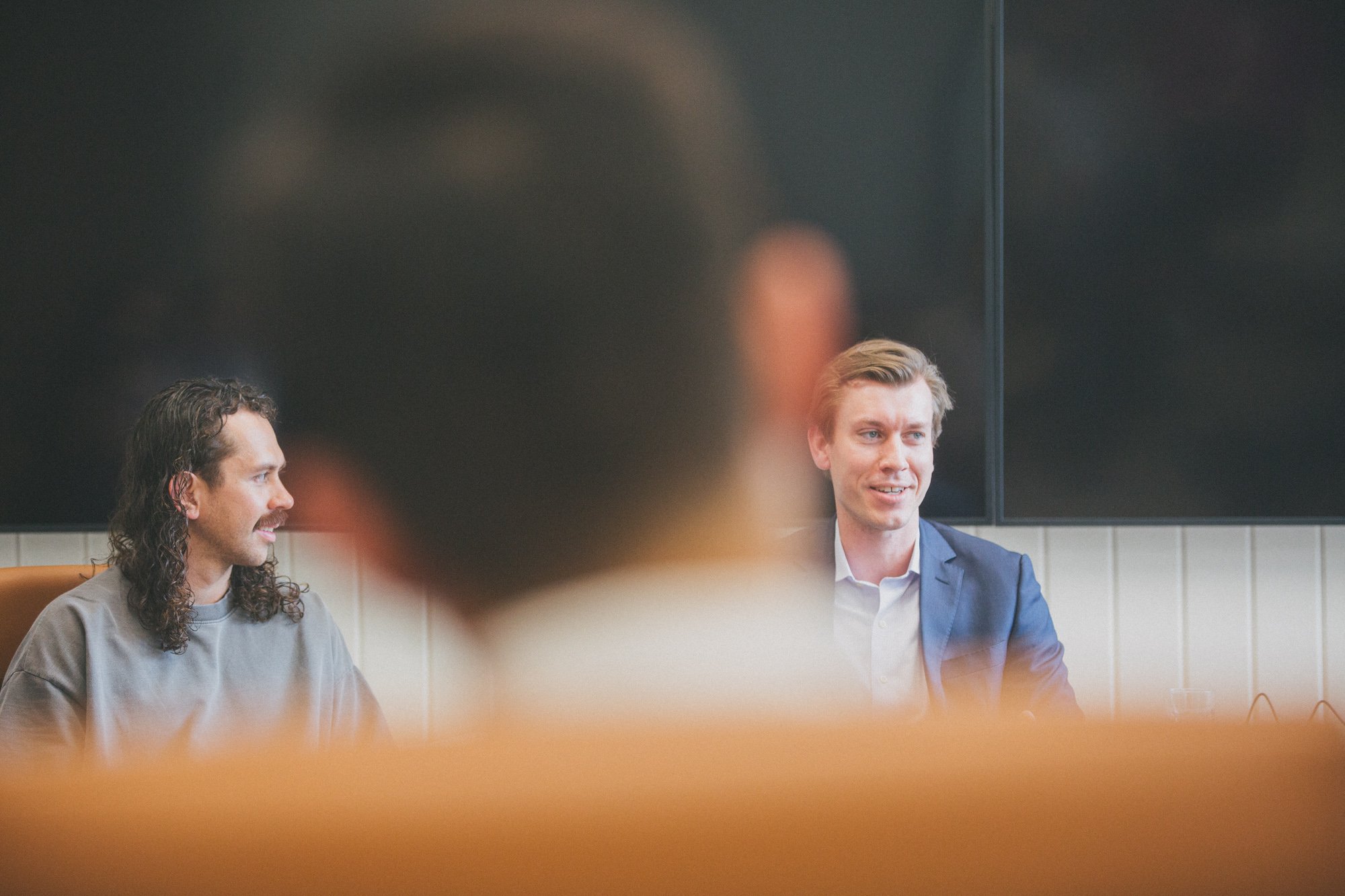Two men are sitting at a table in a meeting room, engaged in conversation. One man has long curly hair and a mustache, and the other is clean-shaven with short, neatly styled hair. A large black screen is behind them.
