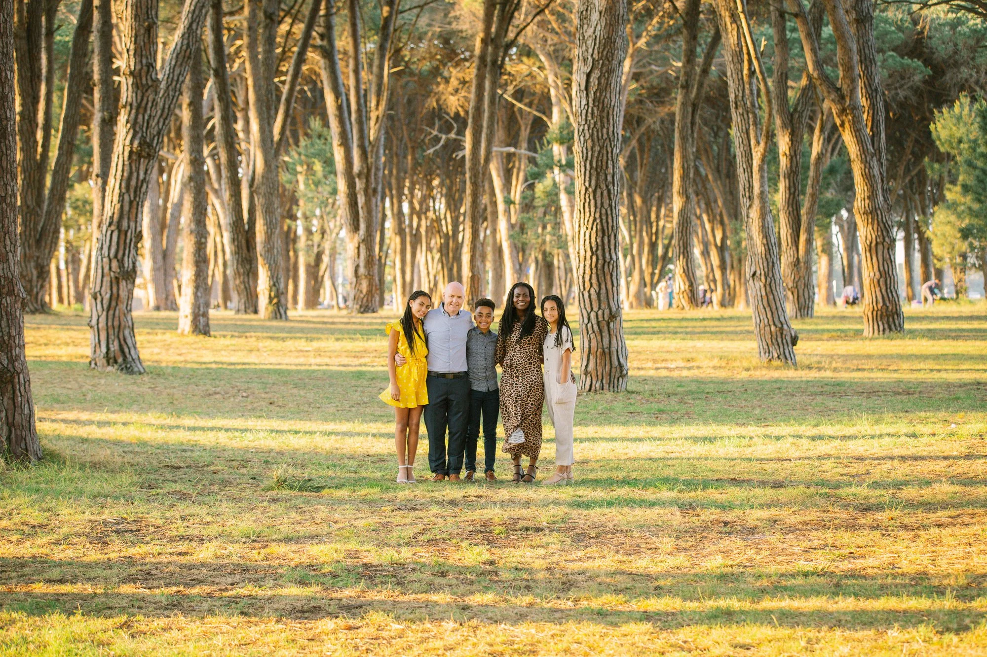 A diverse group of five people standing together in a forest with tall trees, smiling at the camera during sunset.