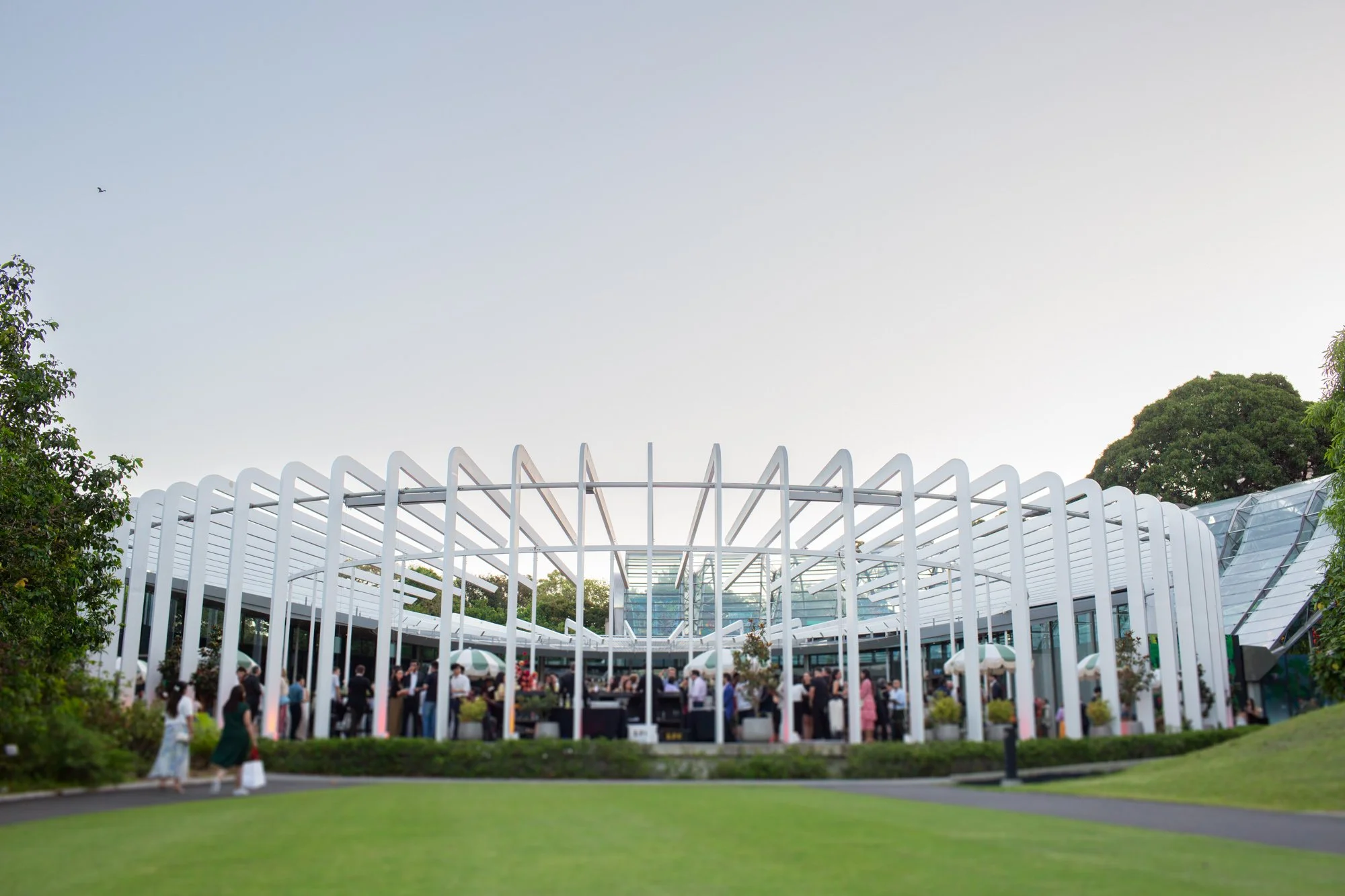 A modern event space with white curved vertical beams, glass walls, and a crowd of people gathered outside during the daytime.