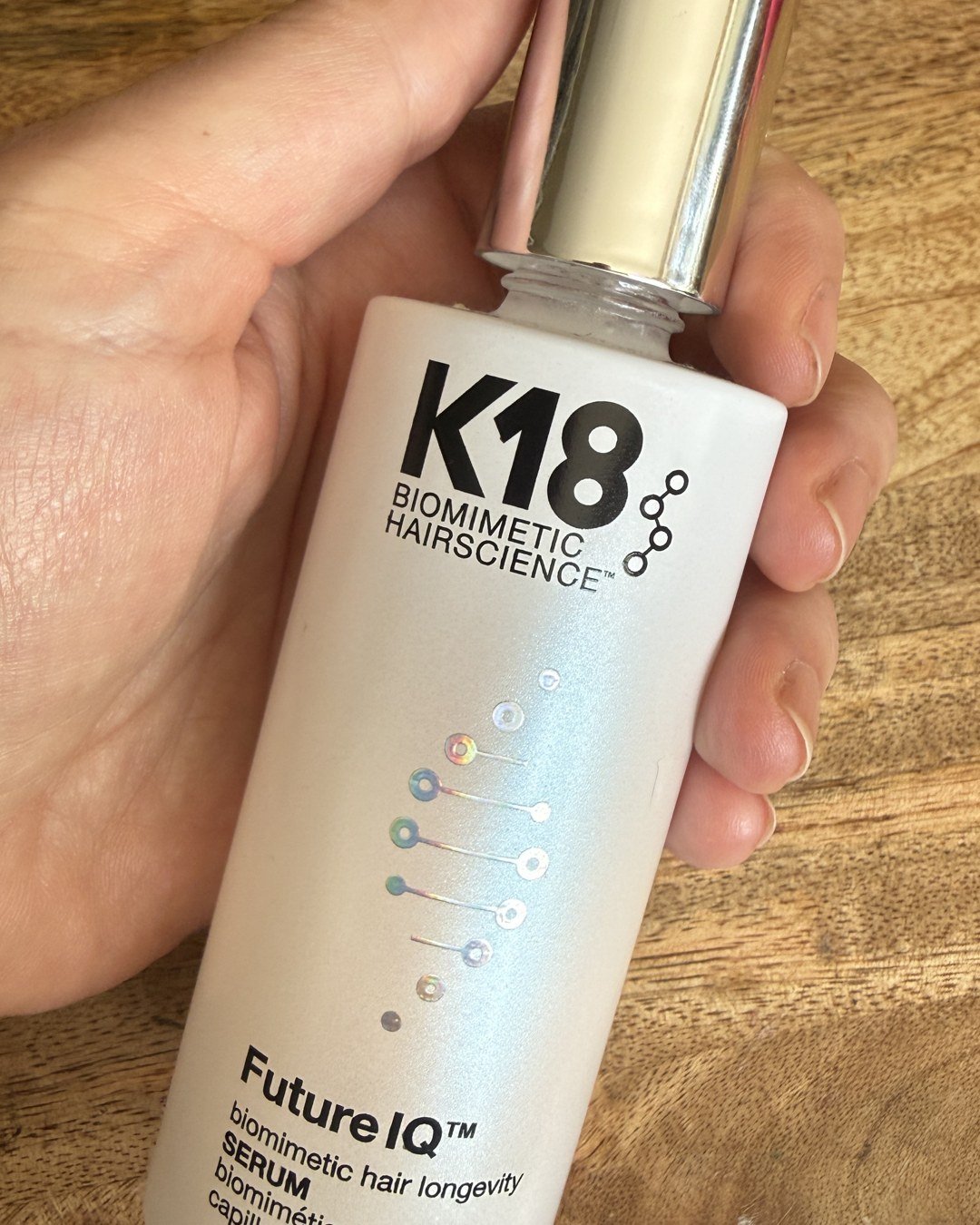 Hand holding a bottle of K18 FutureIQ hair longevity serum over a wooden counter
