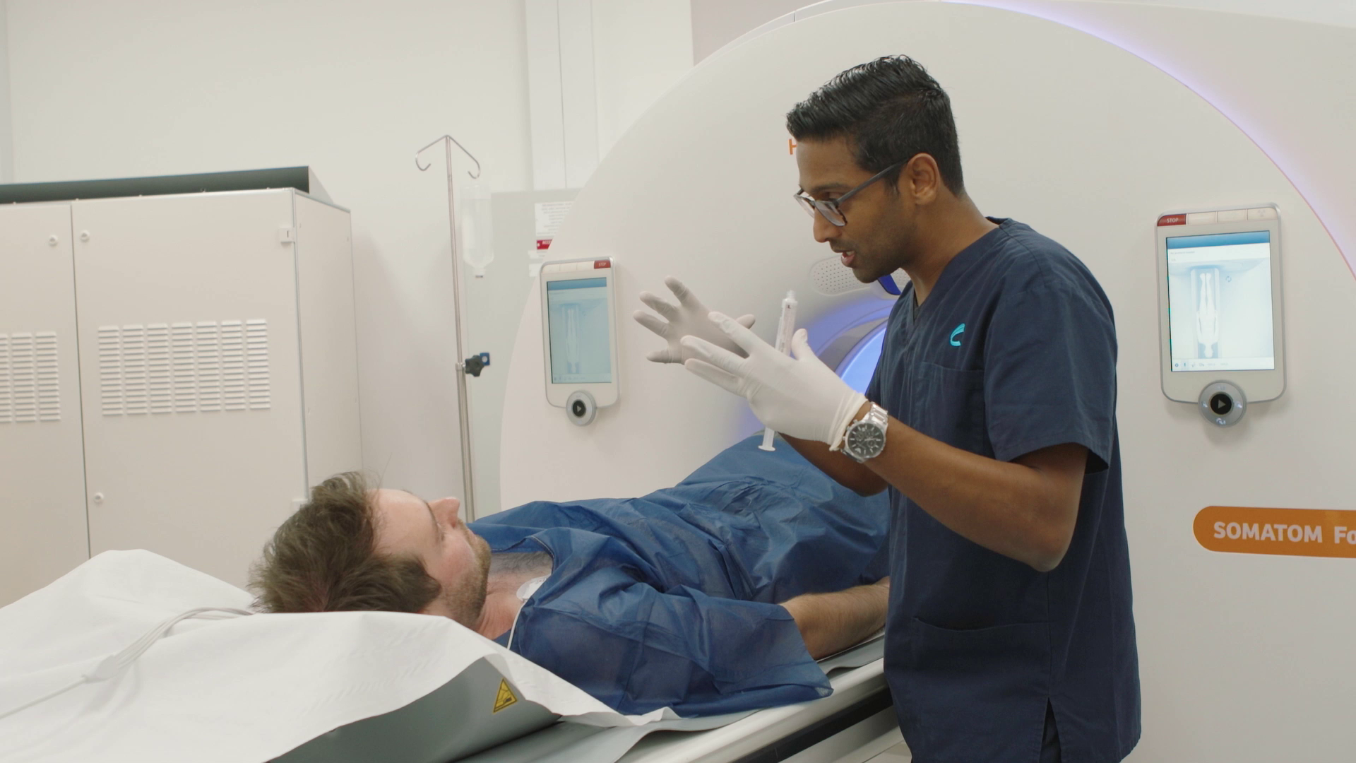 A medical professional preparing to operate an MRI machine while a male patient lies on the MRI table. The professional is wearing gloves and a navy scrubs, and there are monitors on the MRI machine displaying images.