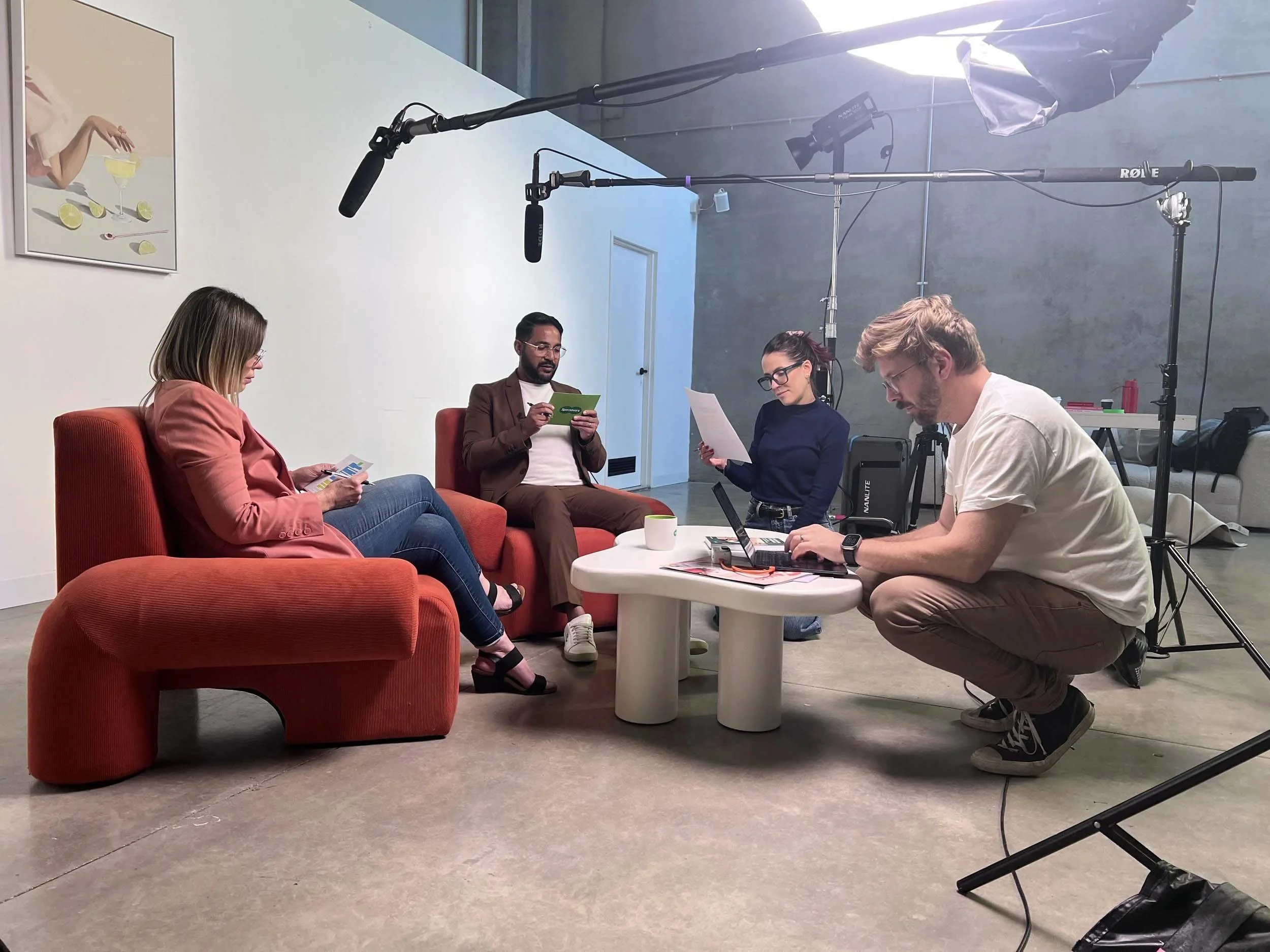 A group of four people seated and working on laptops or reading papers during a video recording or interview session in a studio with professional lighting and camera equipment.