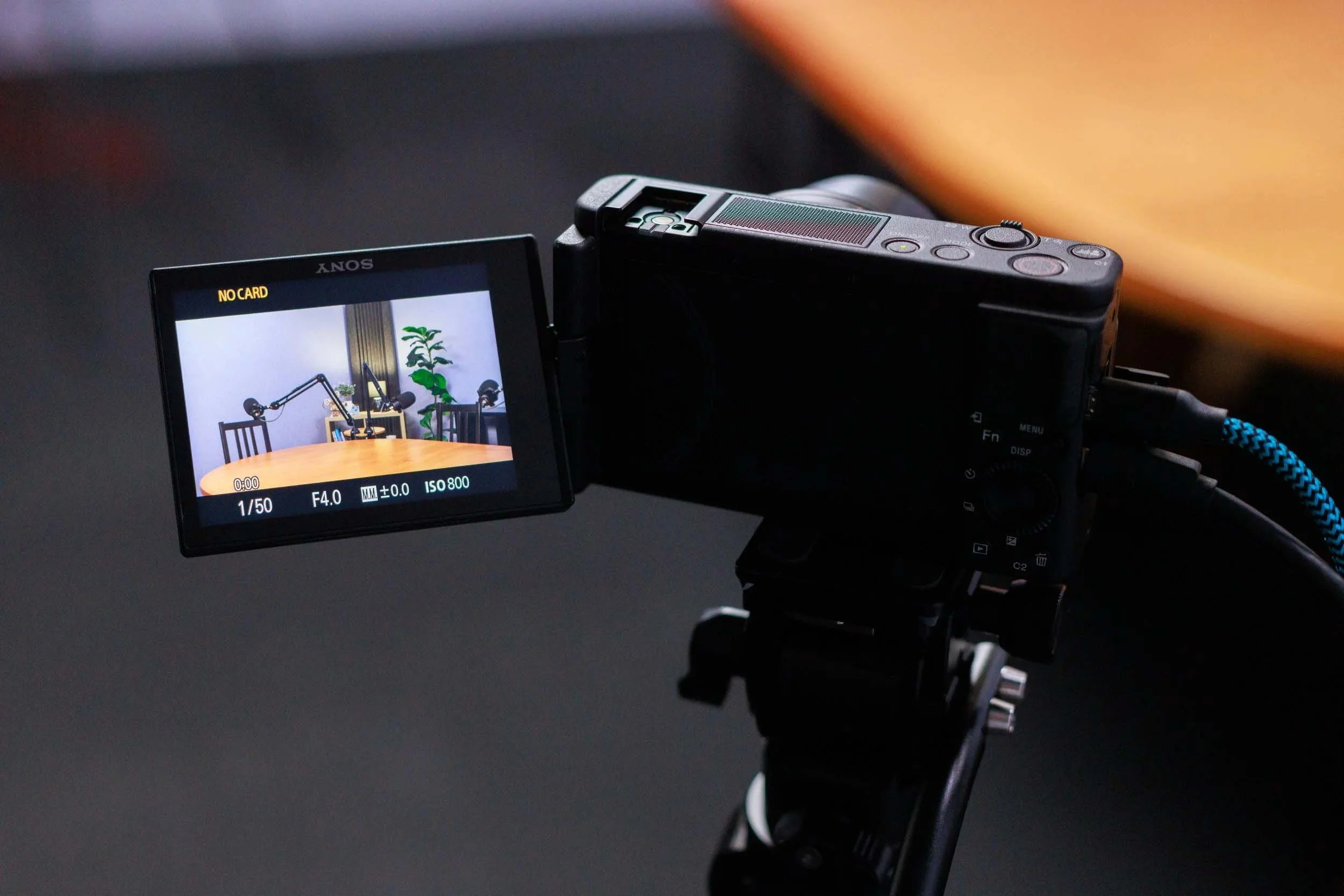 Close-up of a digital camera with a flip-out screen showing a recording of a podcast setup, including microphones and a plant in the background.