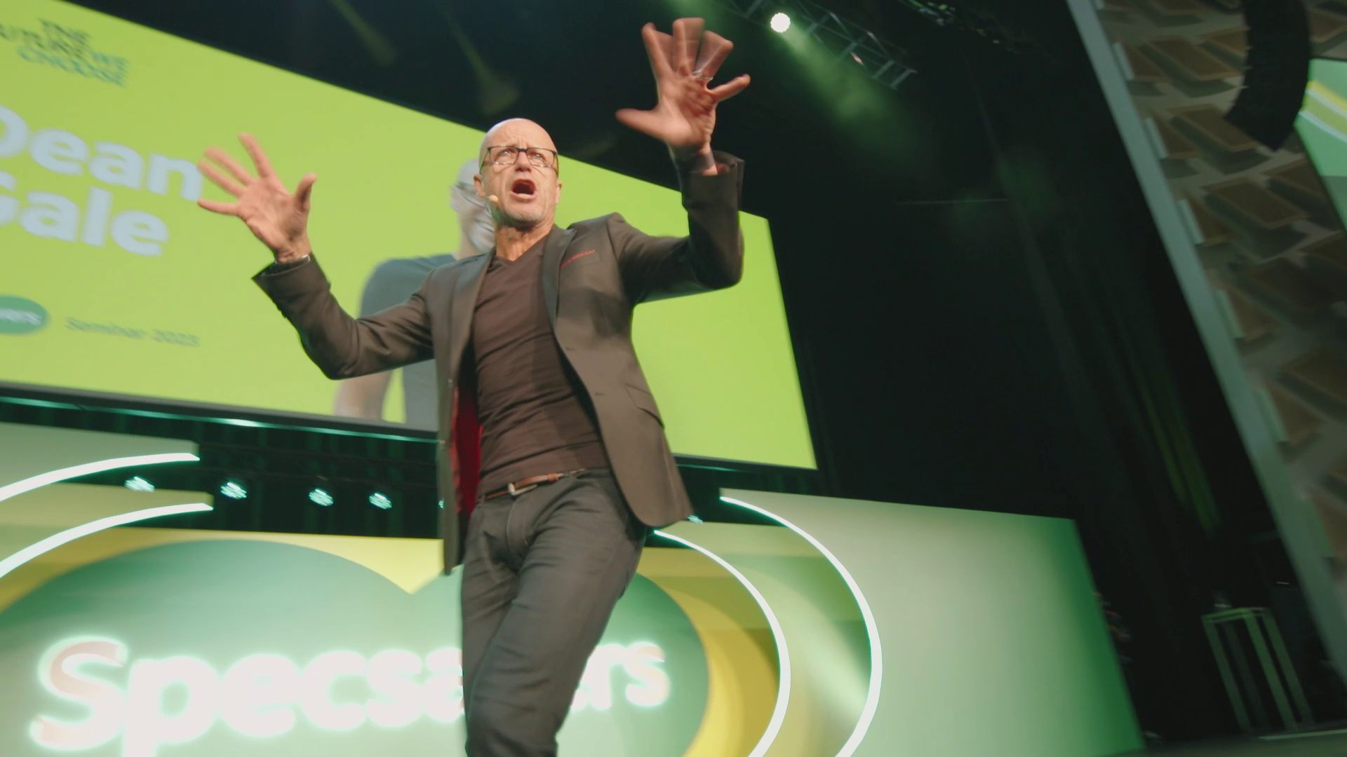A man with glasses and a black blazer, standing on stage at a presentation or event, gesturing with his hands and speaking, with a large yellow-green screen behind him displaying the words 'Specials' and some other text.