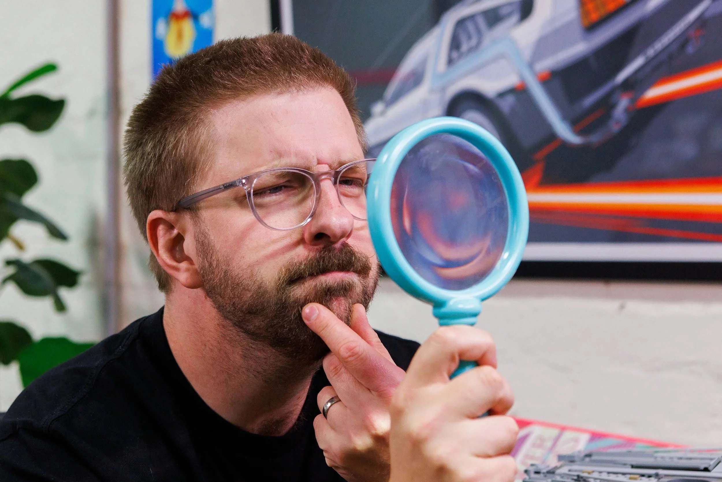 A bearded man with glasses holds a blue magnifying glass close to his eye, appearing to observe something with curiosity in an indoor setting, with a colorful racing car poster on the wall behind him.