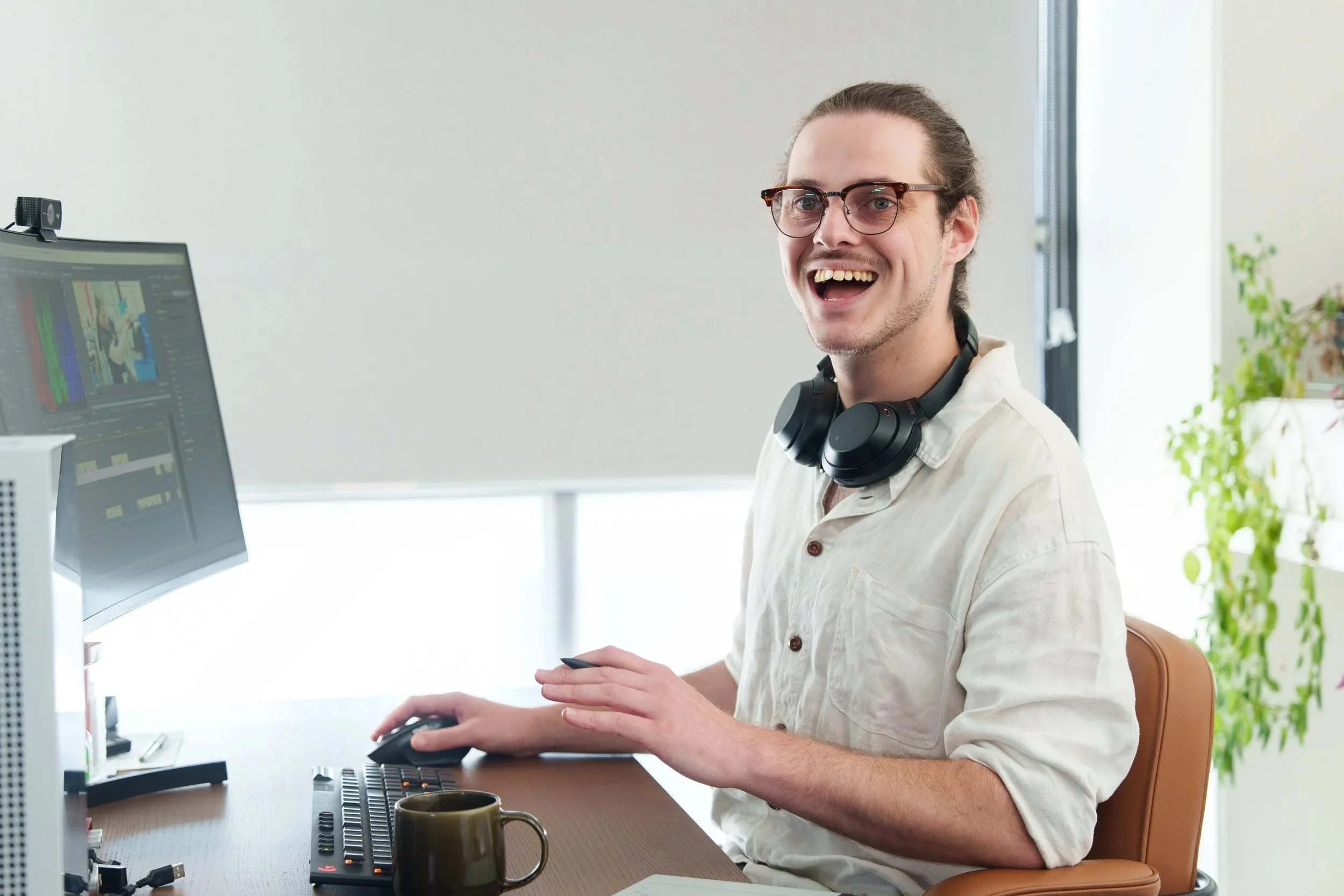 A man with glasses, a light-colored shirt, and headphones around his neck is smiling while working at a desk with a computer, in a bright room with a window and a green plant in the background.